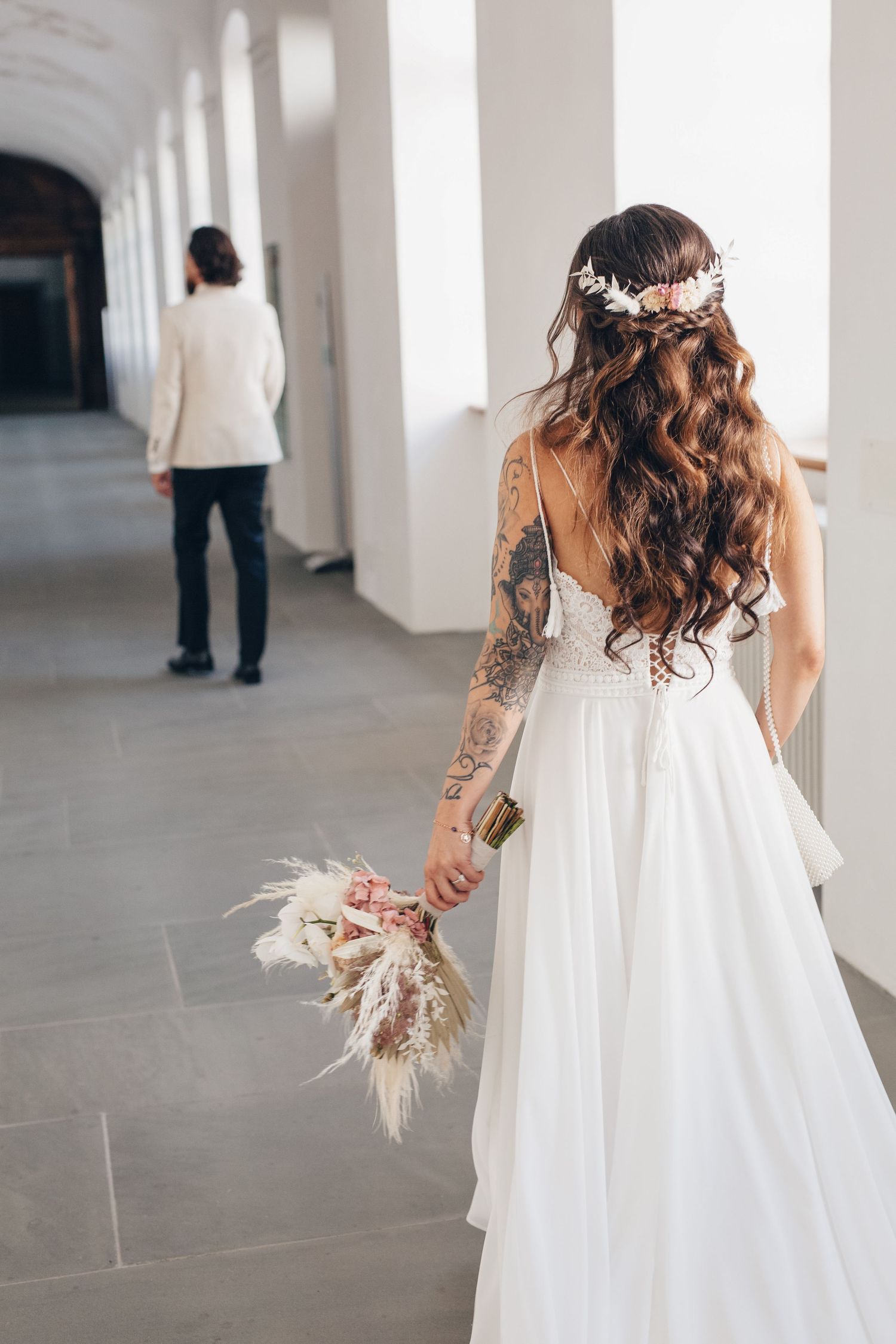 Bride in white gown with floral crown holds bouquet, facing groom in white suit down elegant hallway.