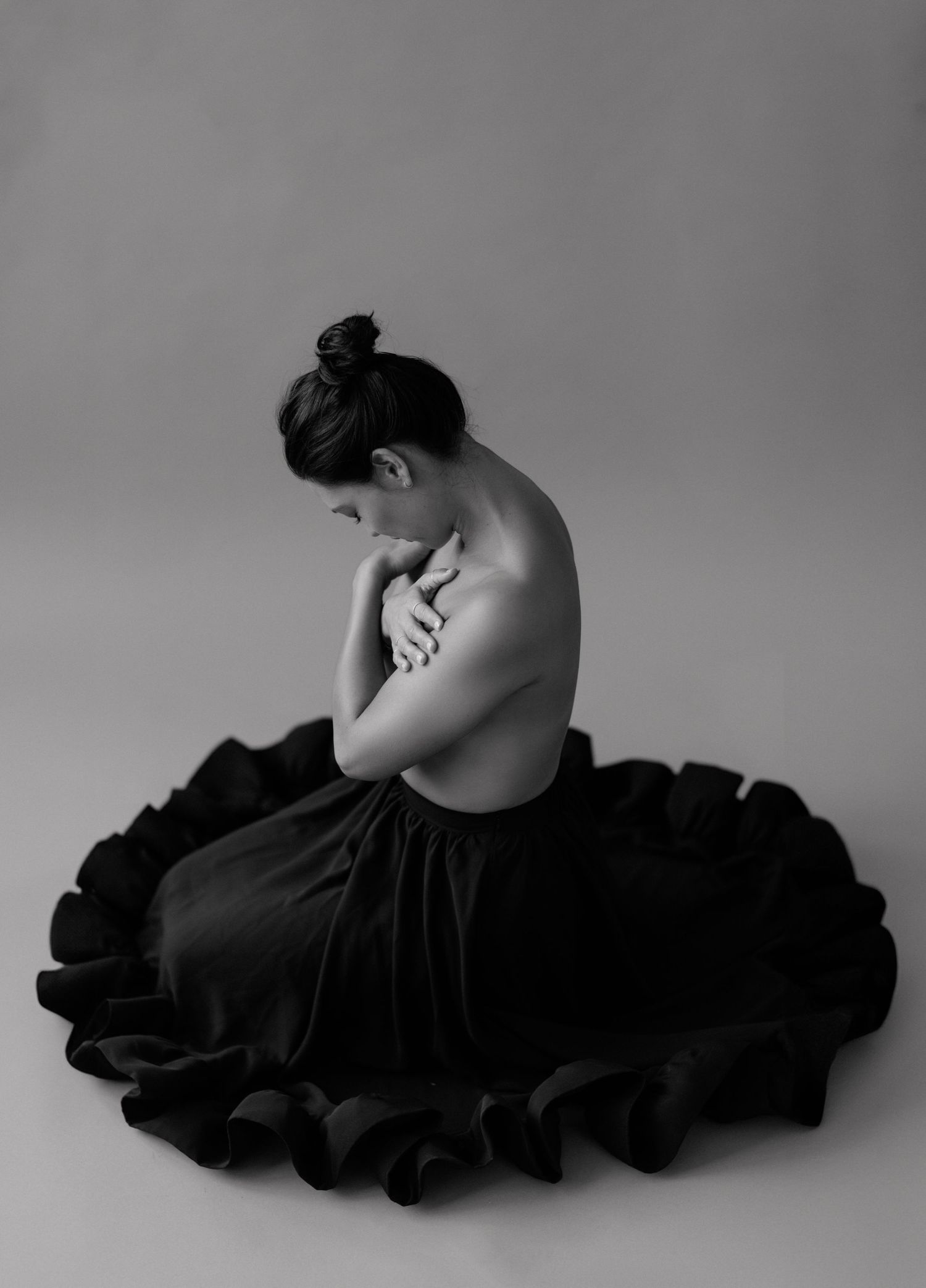 Black and white photo of a woman in a dark ruffled skirt, seated with arms crossed, head bowed in a graceful pose.