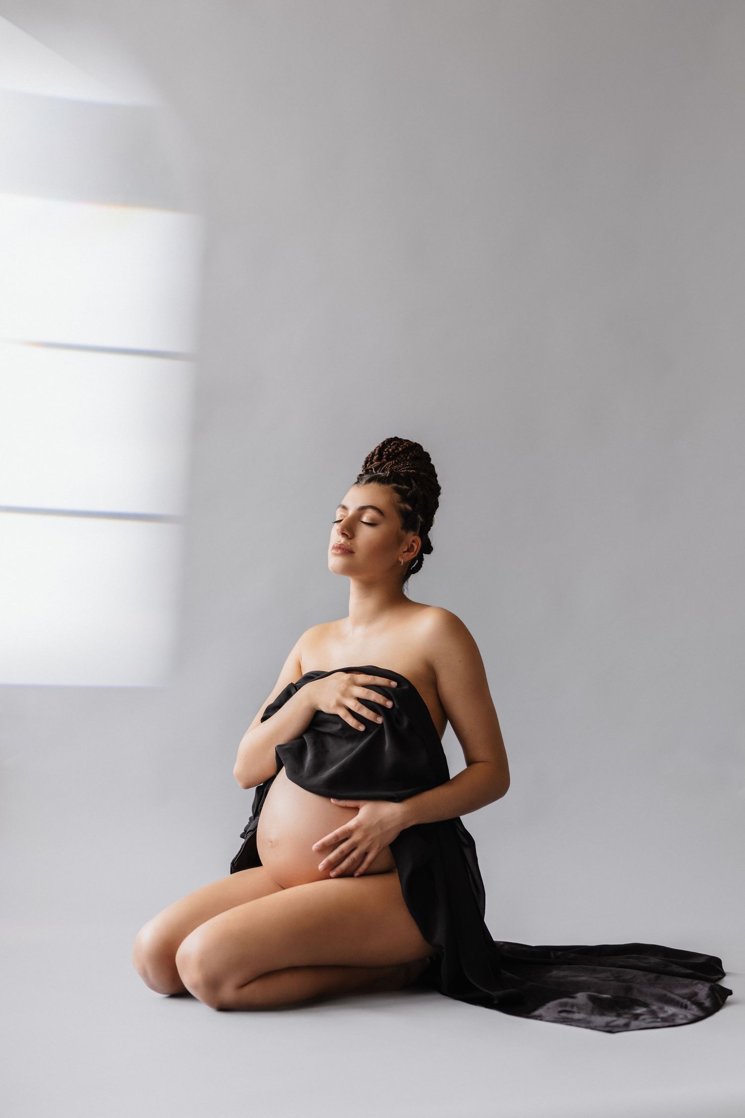 Pregnant woman sitting gracefully on floor, draped in black fabric, cradling her baby bump in a serene studio setting.