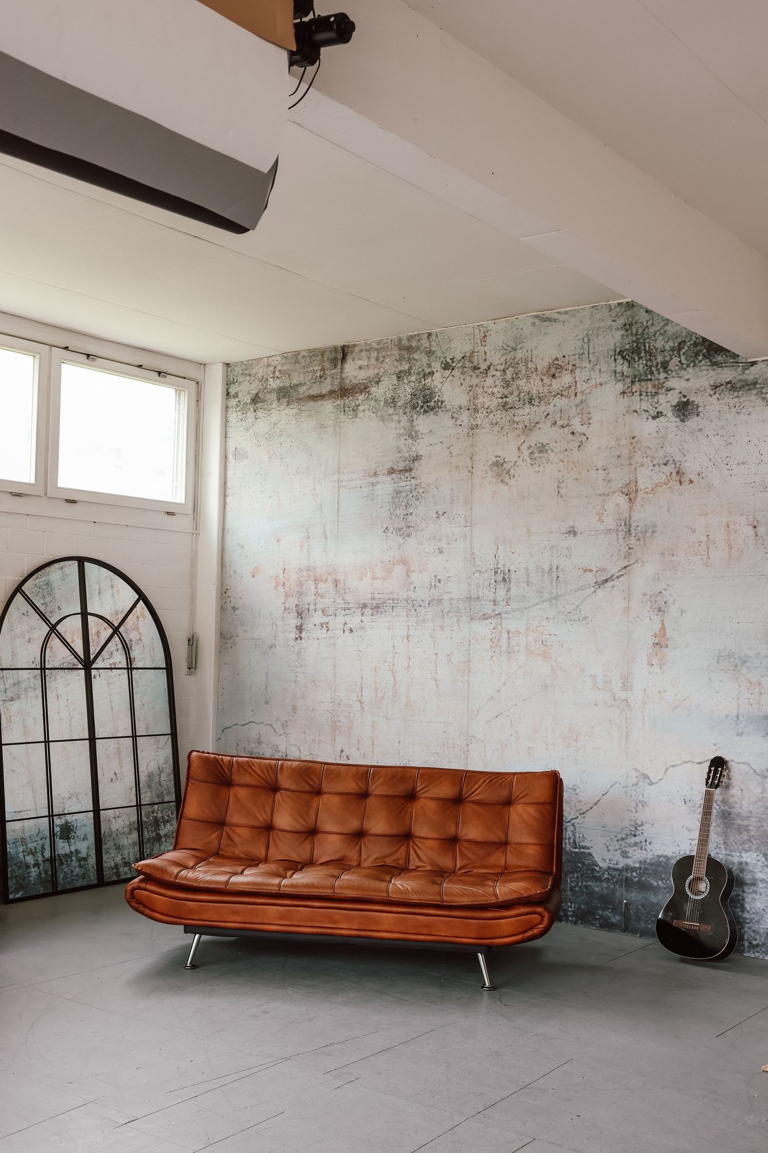Vintage brown leather tufted sofa against distressed wall with acoustic guitar and arched mirror in loft space.