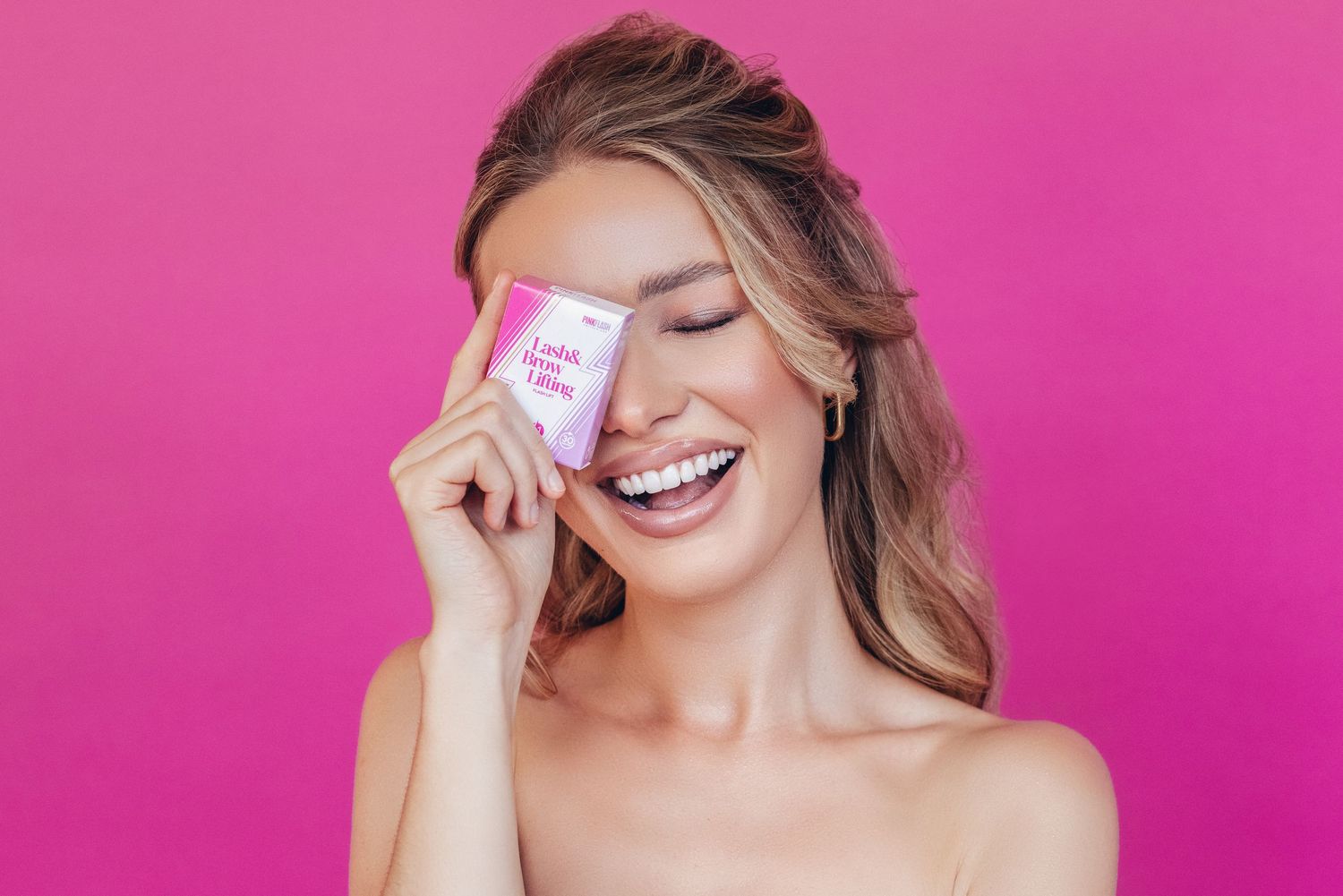 Smiling woman holding a pink product card against a vibrant pink background, radiating joy and beauty.