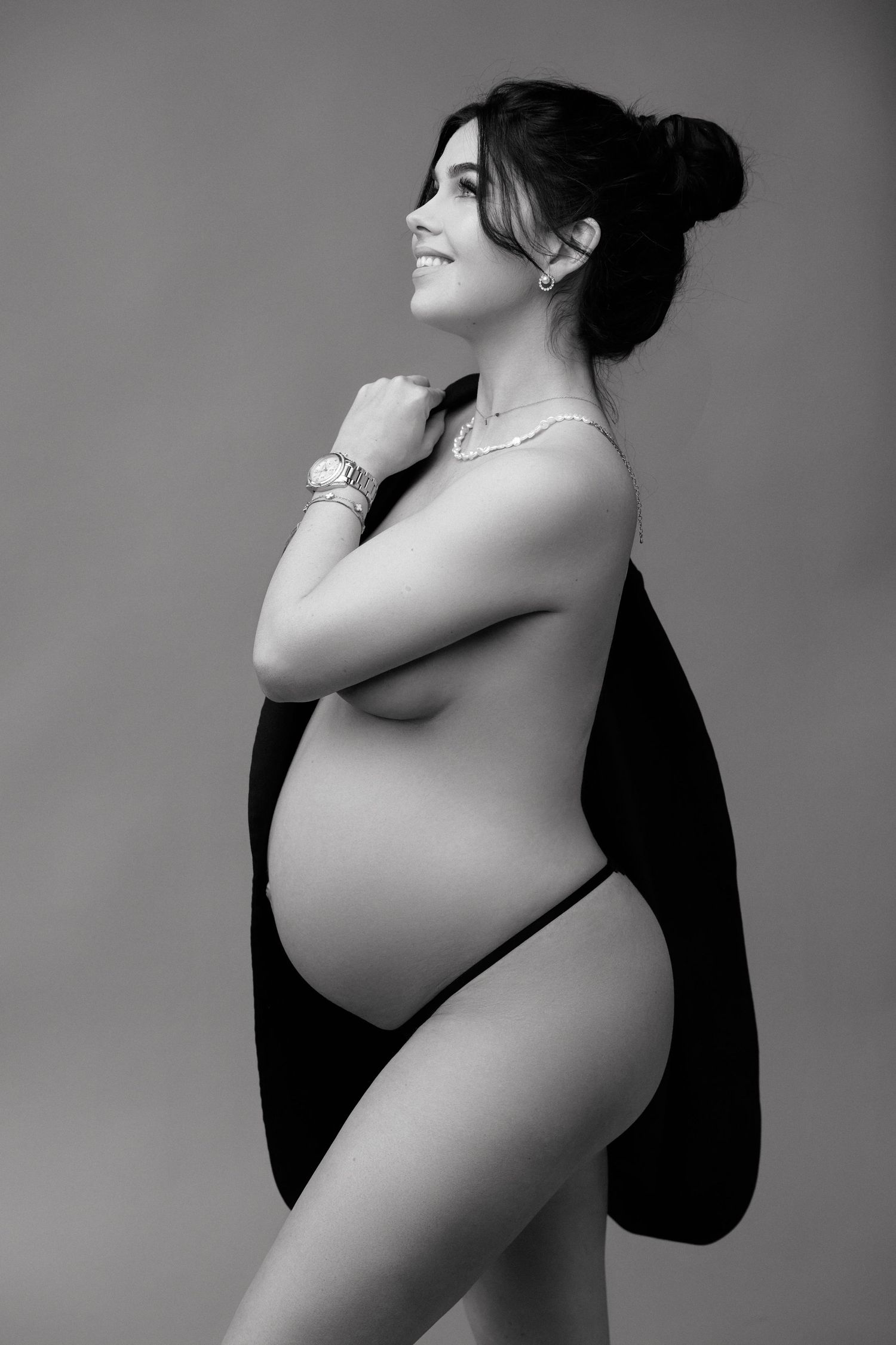 Black and white maternity photo of smiling pregnant woman posing sideways, wearing jewelry and draped fabric.