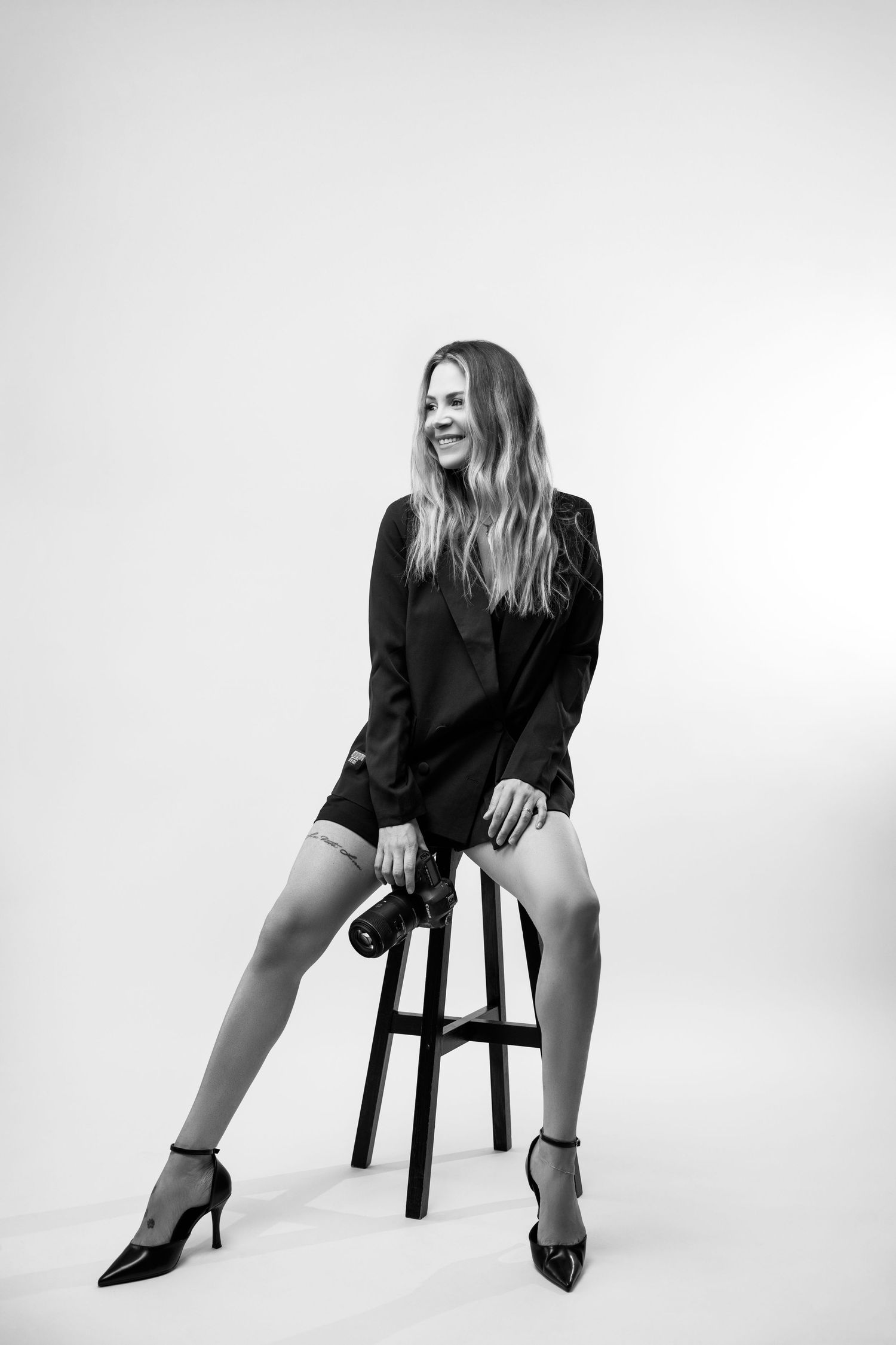 Black and white photo of a smiling blonde woman in black blazer and heels sitting on a stool holding a camera.