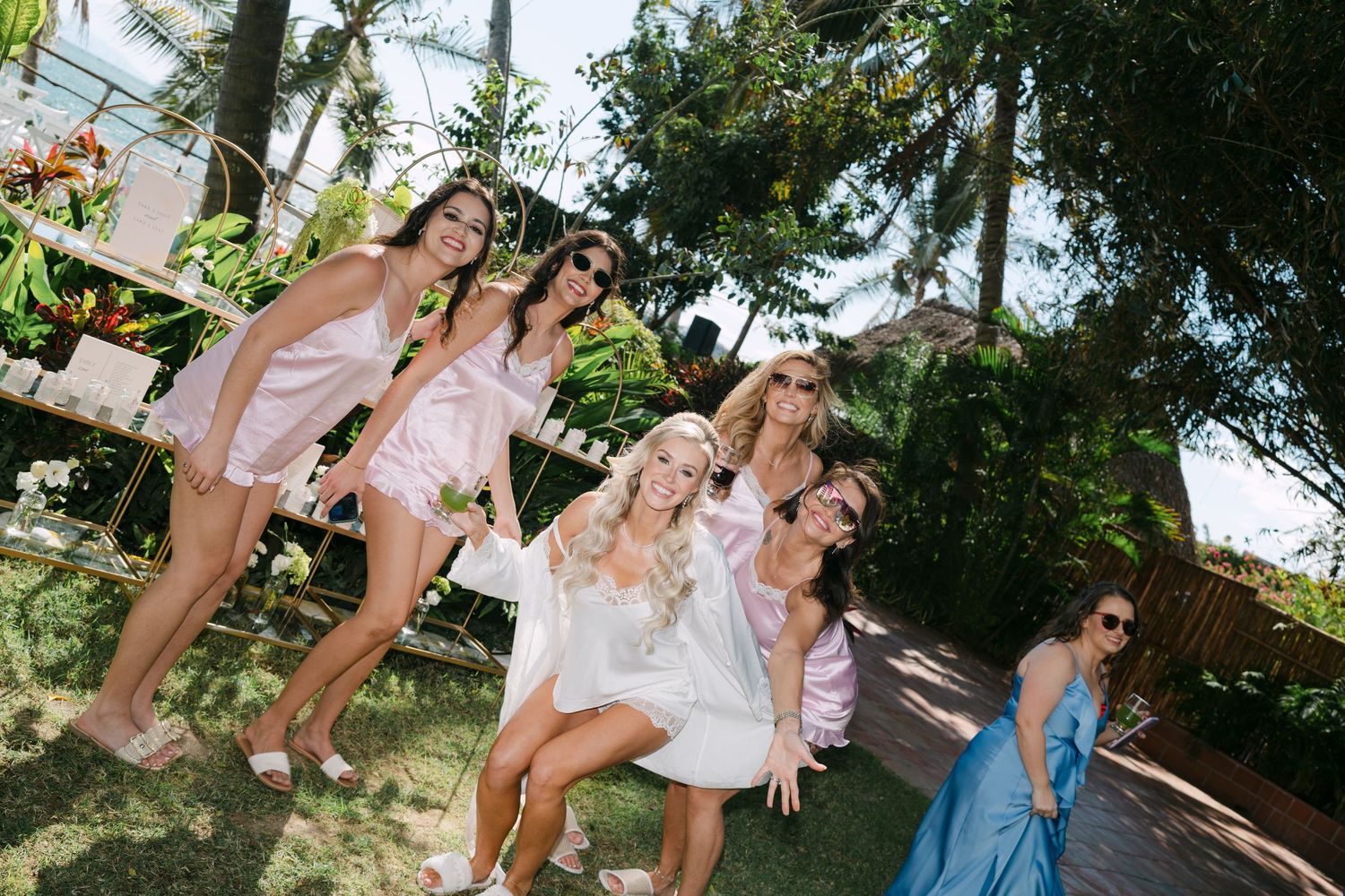 Group of bridesmaids in pink and white outfits laughing and posing together in a tropical garden setting.