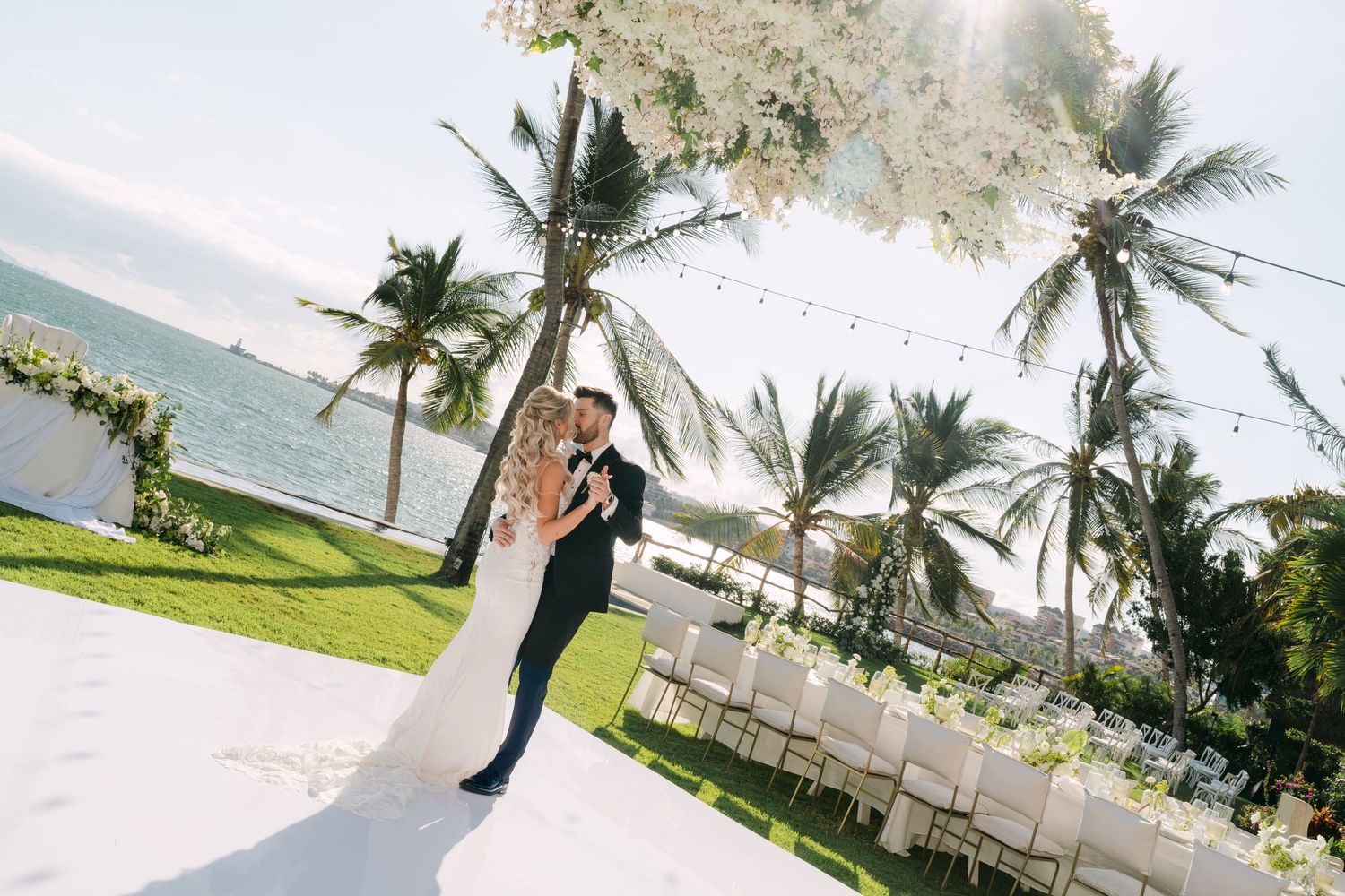 Bride and groom sharing first dance under floral arch with ocean view, palm trees and string lights at sunset.