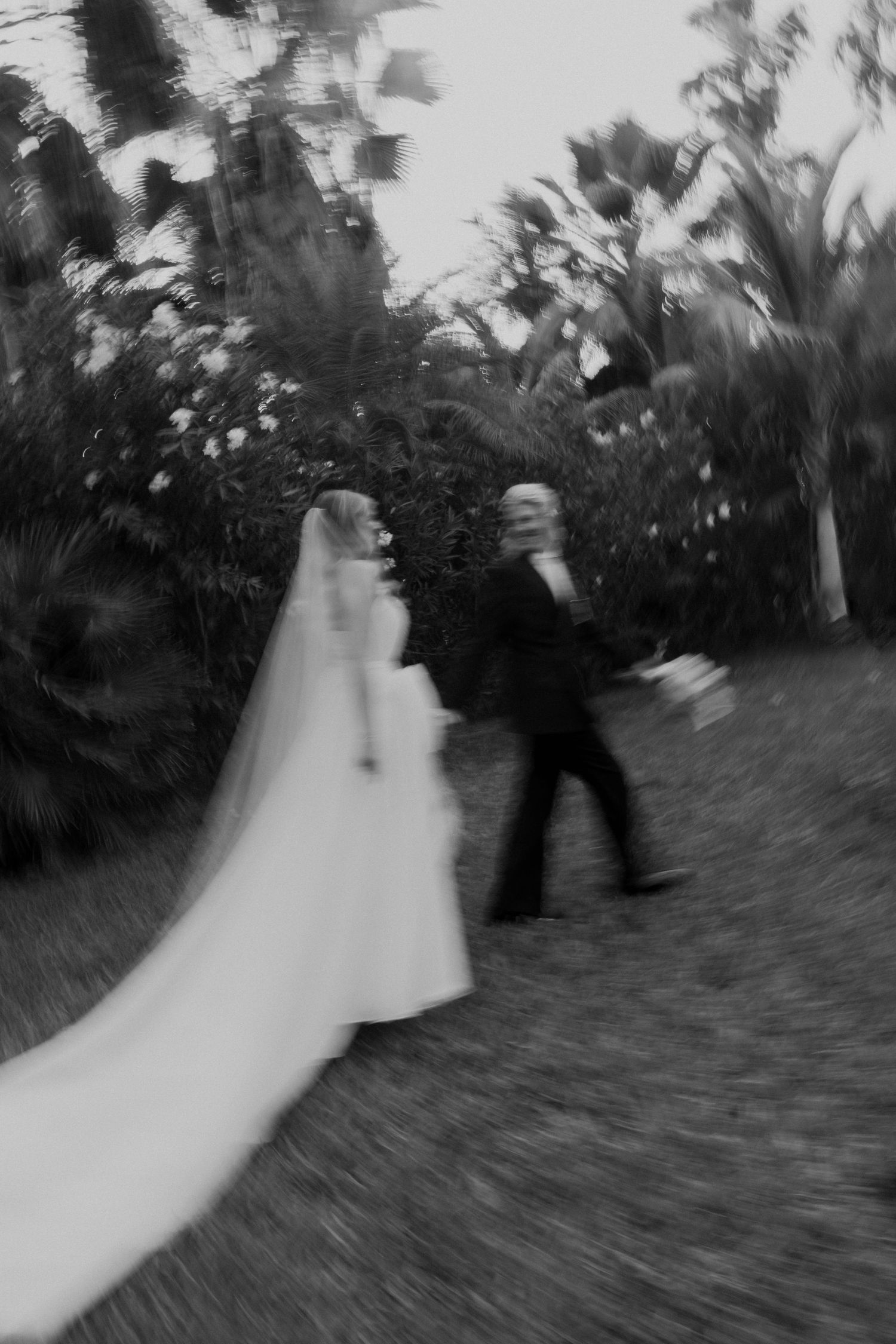 Black and white blurred photo of bride and groom walking through lush garden after ceremony.
