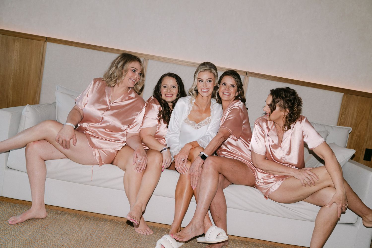 Bride and four bridesmaids in matching pink satin robes sitting together on a white sofa laughing and smiling.