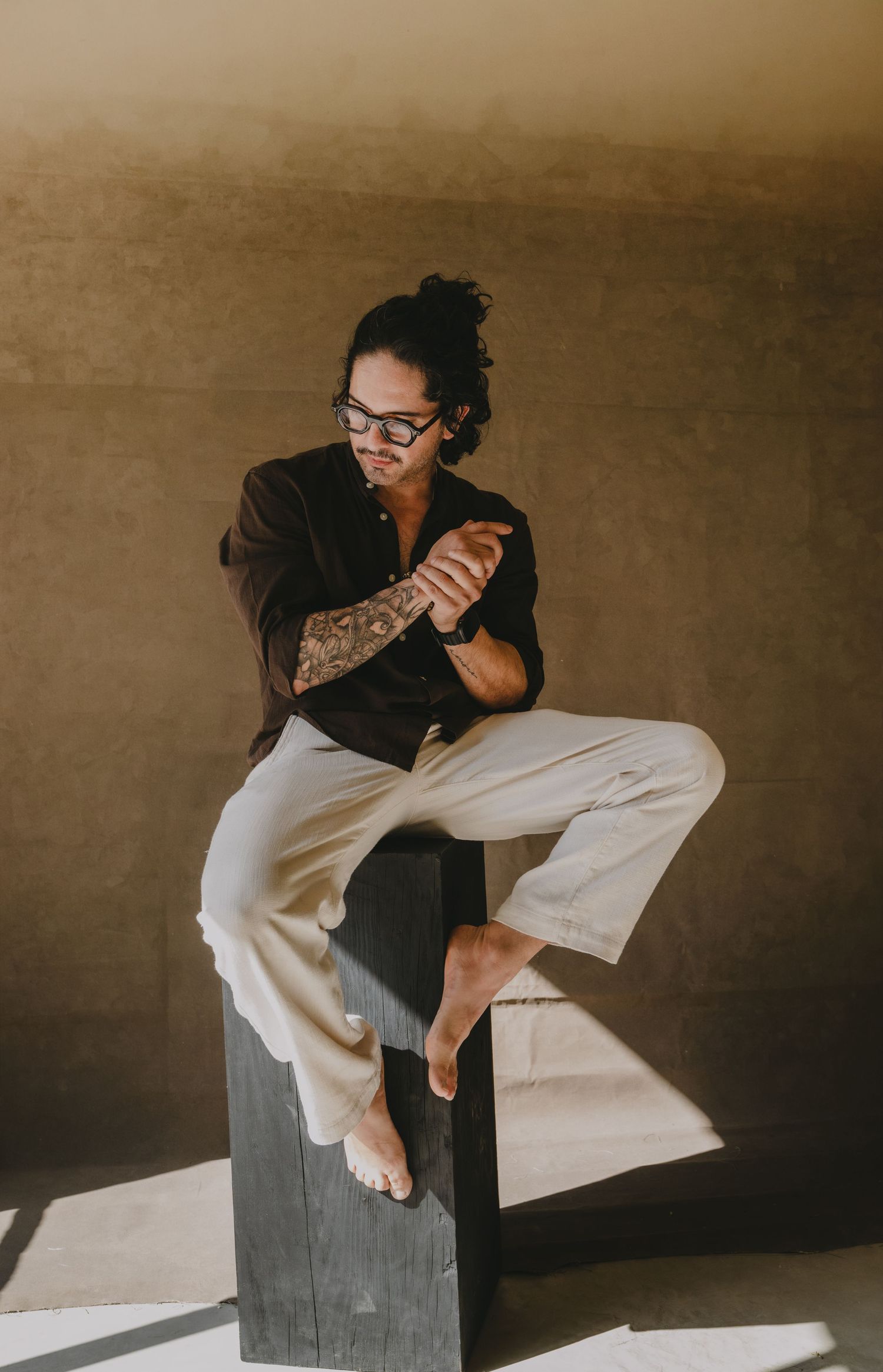 Tattooed person with glasses in black shirt and beige pants sitting barefoot on dark wooden post indoors.
