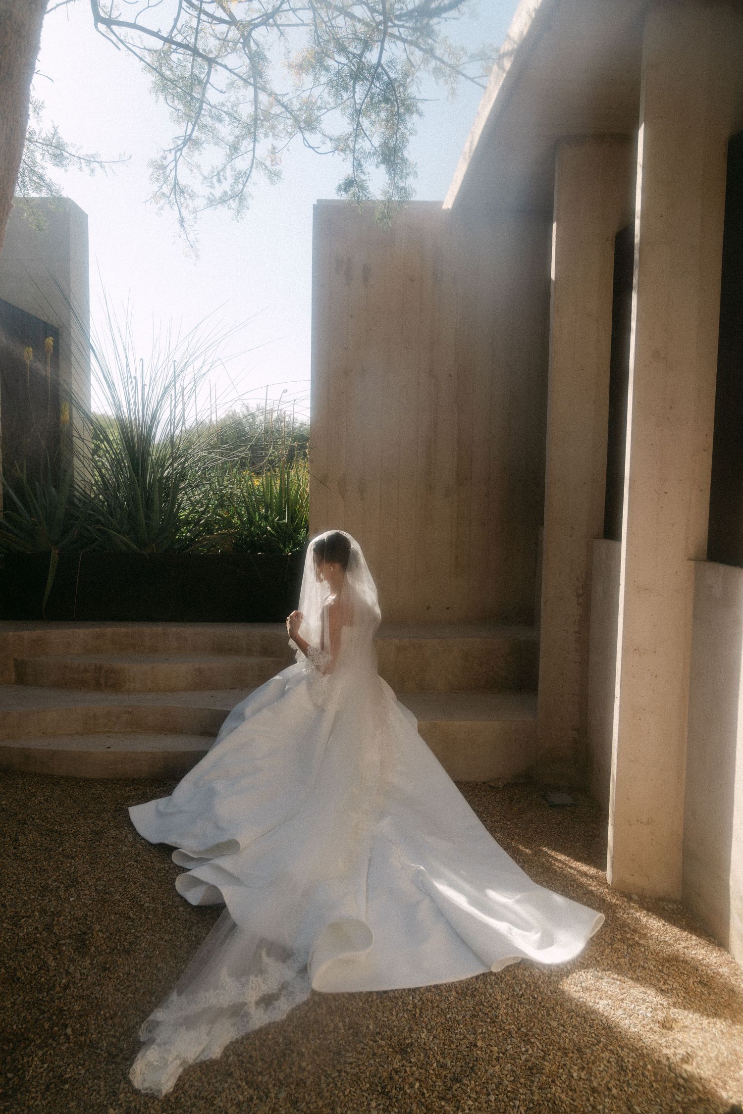 Bride in a voluminous white wedding gown with veil stands between concrete columns in sunlight.