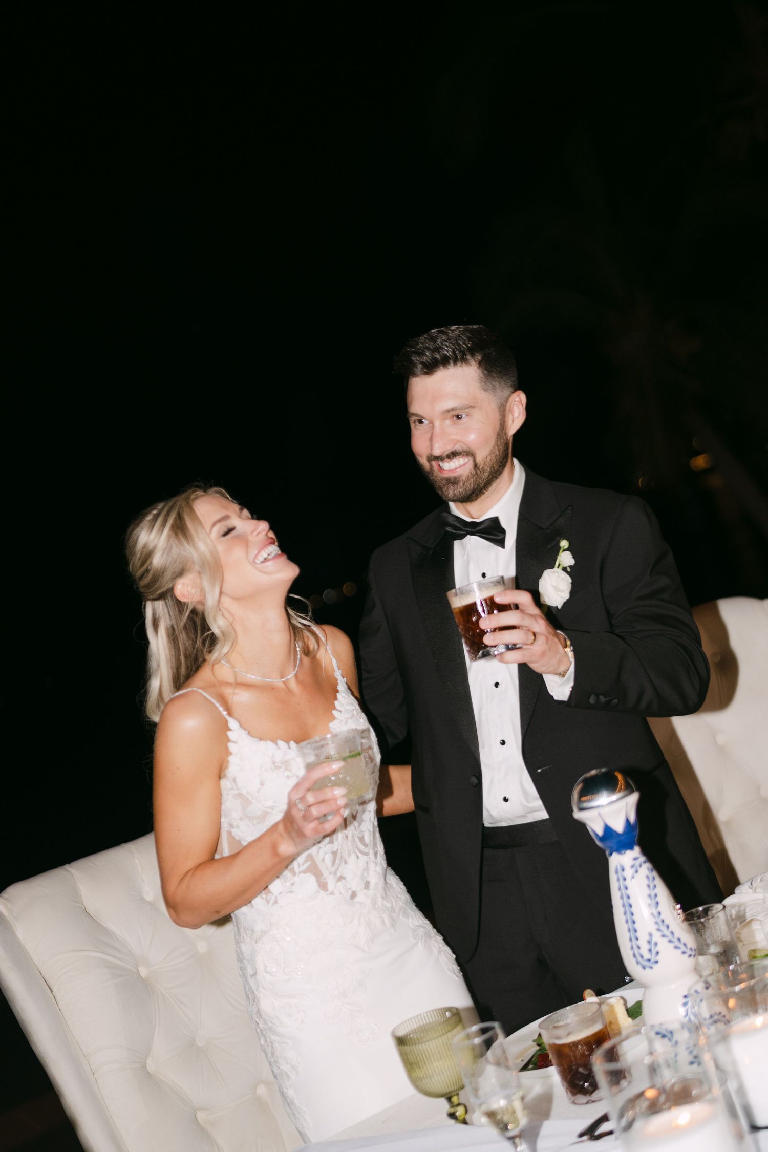 Bride and groom laugh and raise their glasses during the outdoor evening wedding reception.