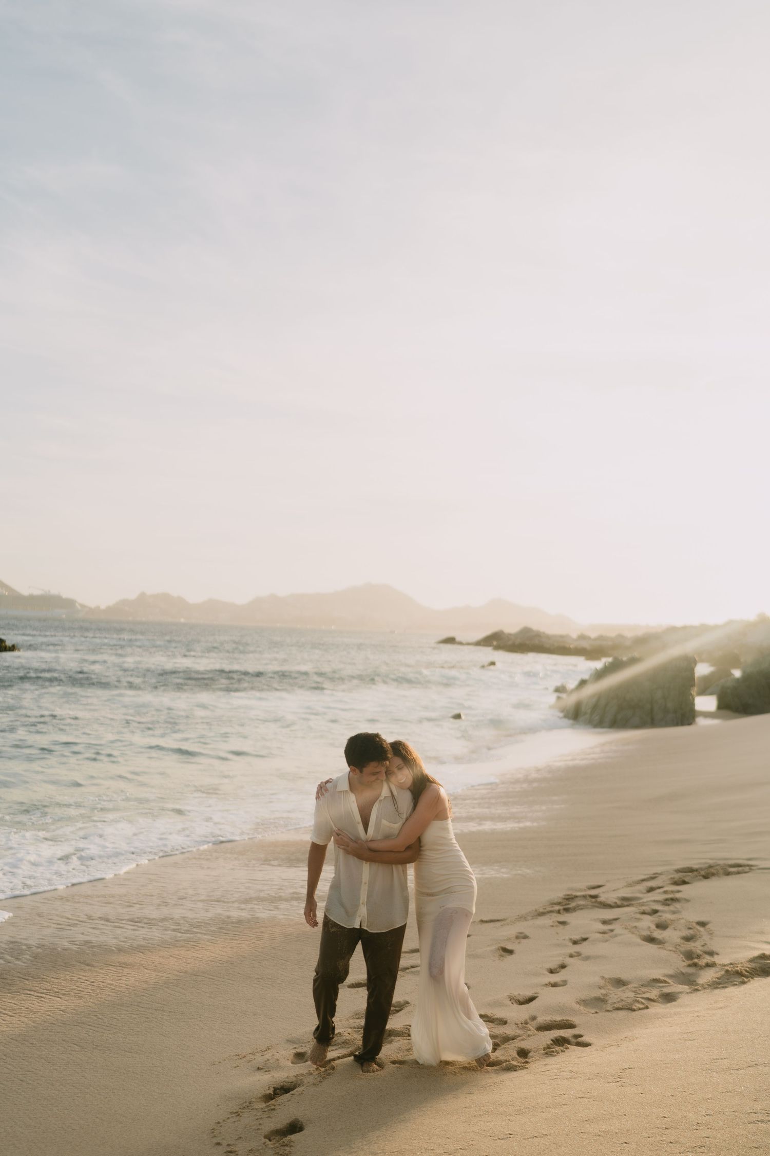 Couple embracing on a golden sandy beach at sunset, waves gently lapping the shore with mountains in the background.