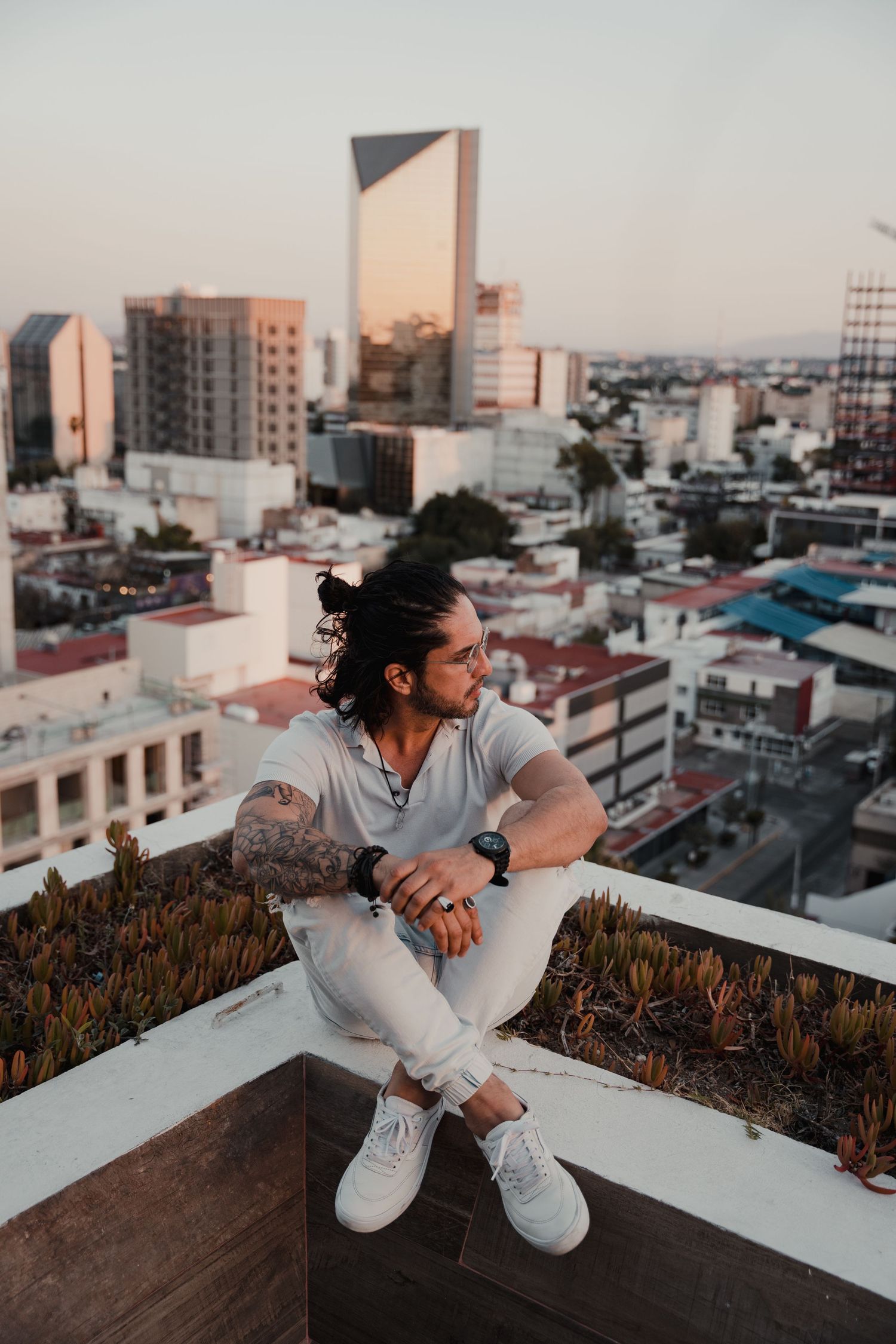 Tattooed man with long hair sitting on rooftop ledge overlooking urban city skyline at sunset.