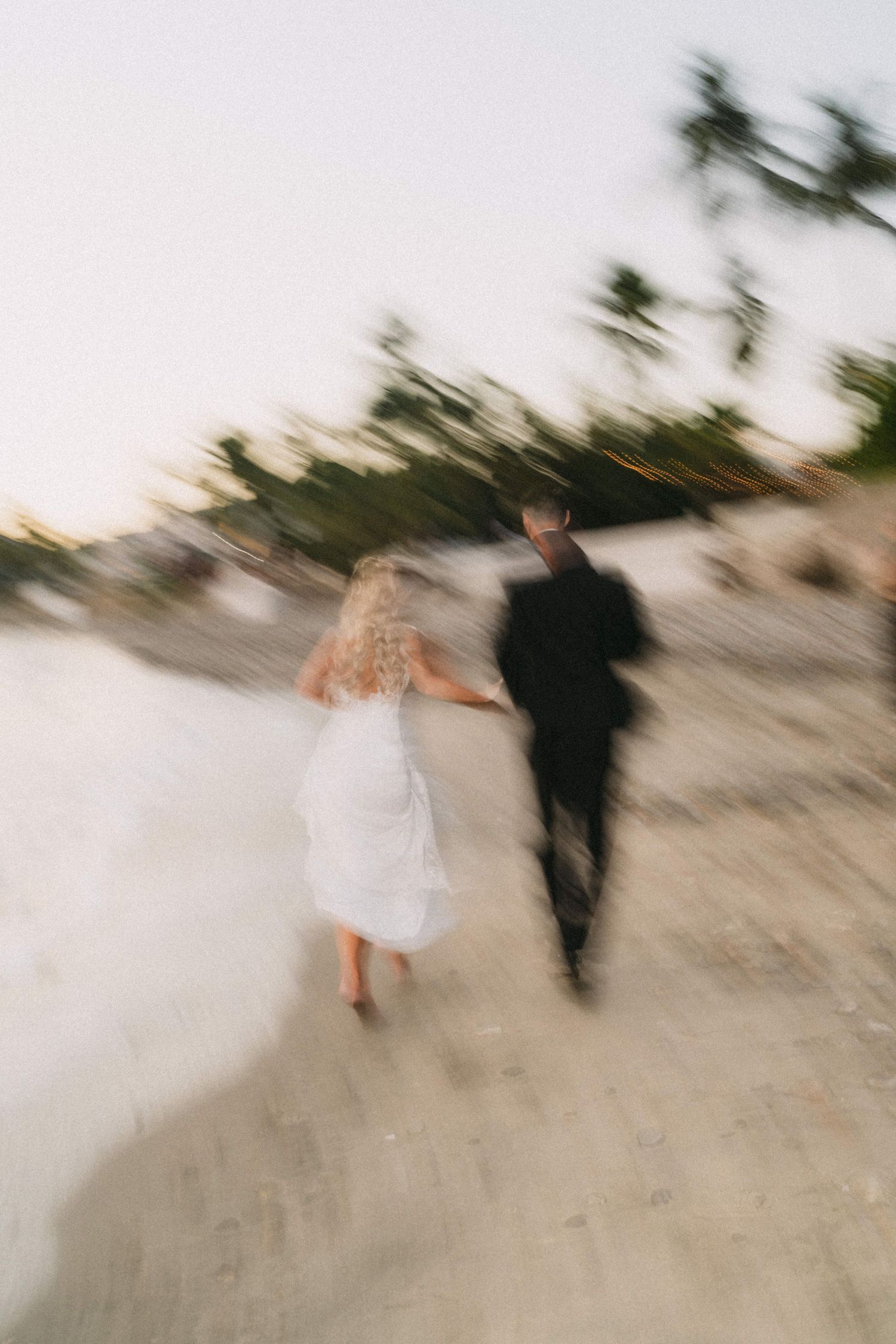 Blurred motion shot of bride and groom holding hands running along a sandy beach at dusk near palm trees.