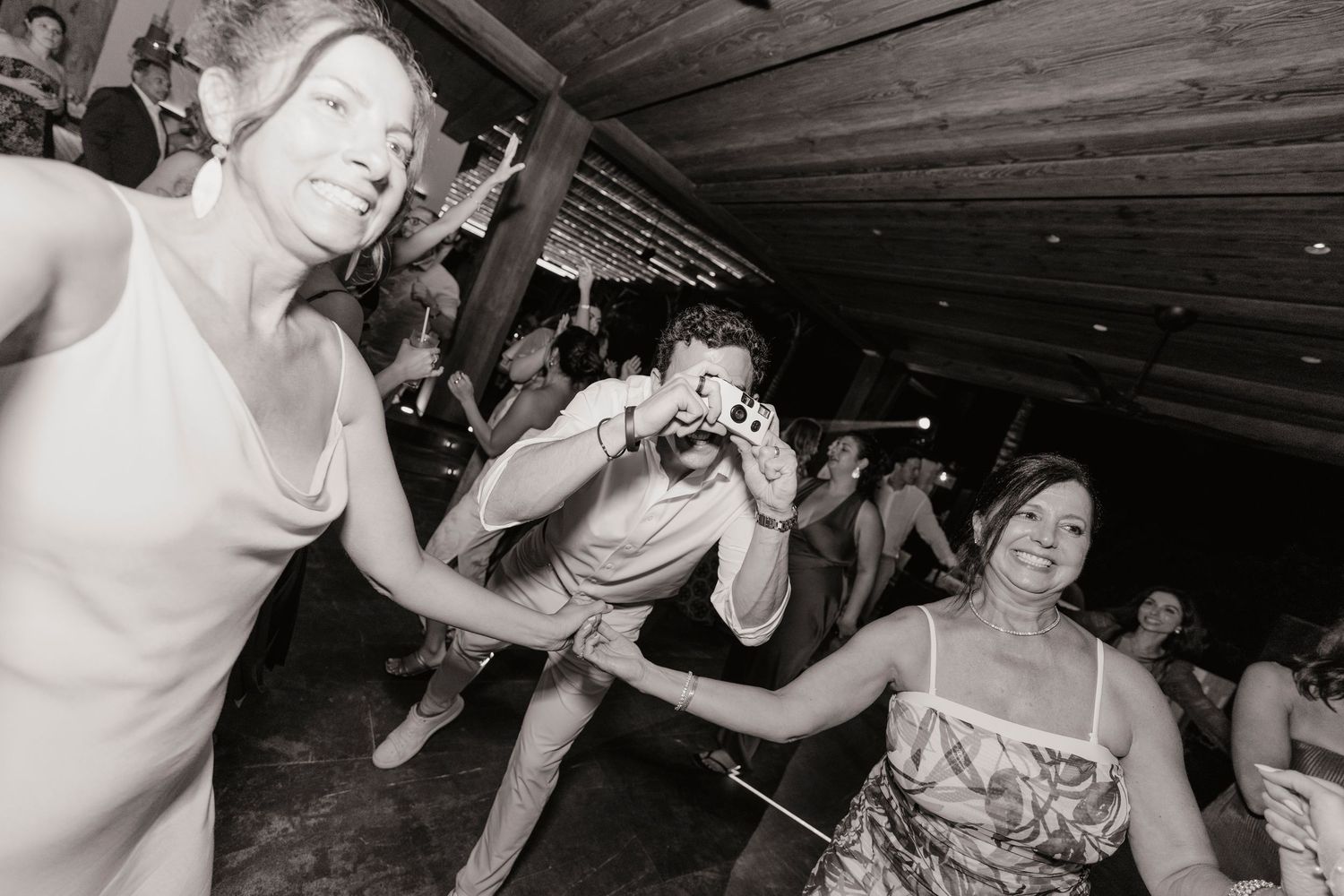 Black and white candid photo of guests dancing and laughing at a lively wedding reception.