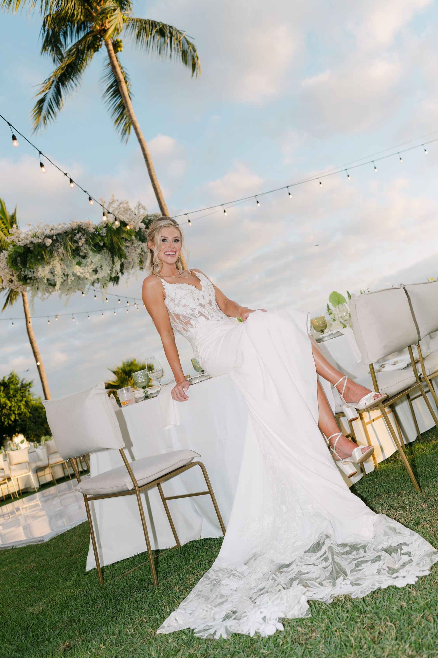 Elegant bride in lace wedding gown posing at outdoor tropical reception with palm trees and string lights.