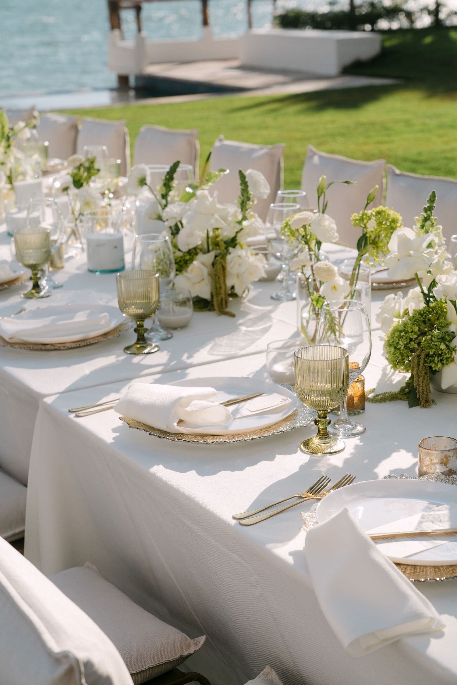 Close-up of elegant wedding reception table with white flowers, sage green glassware, gold flatware, and white linens by the sea.