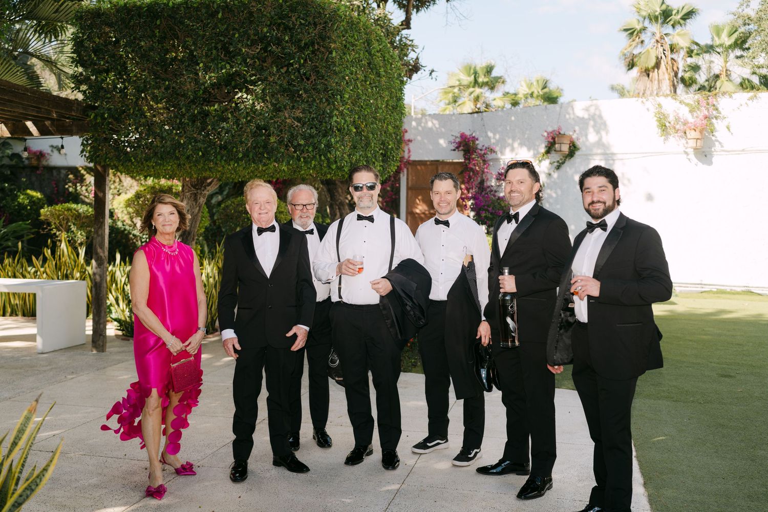 Groomsmen and guests in black tuxedos posing together outdoors with lush greenery and bougainvillea backdrop.