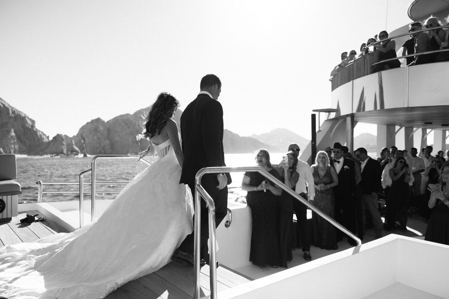 Black and white photo of a bride and groom exchanging vows on a yacht deck with guests and rocky cliffs in background.