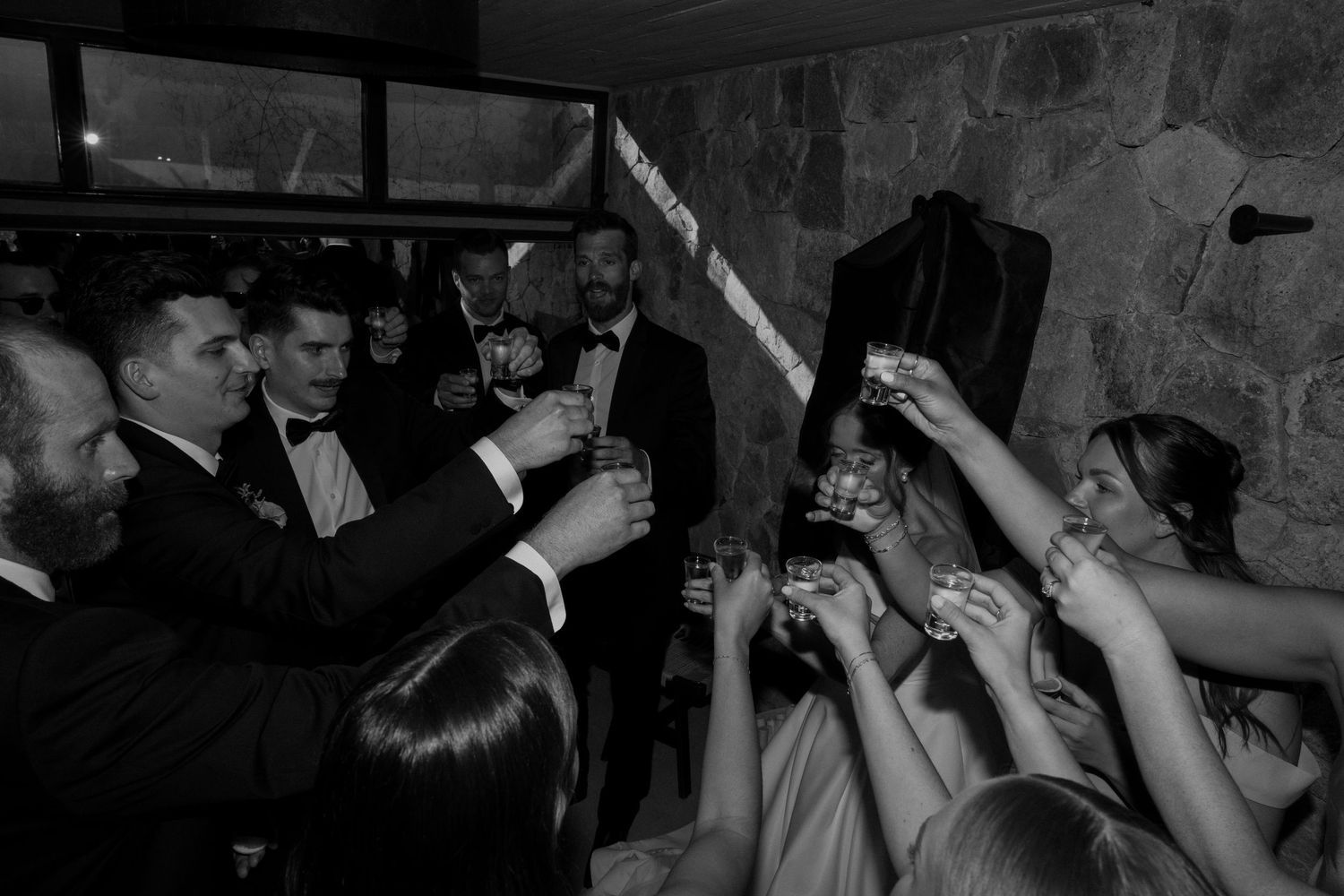Black and white photo of wedding party raising glasses in a celebratory toast indoors.