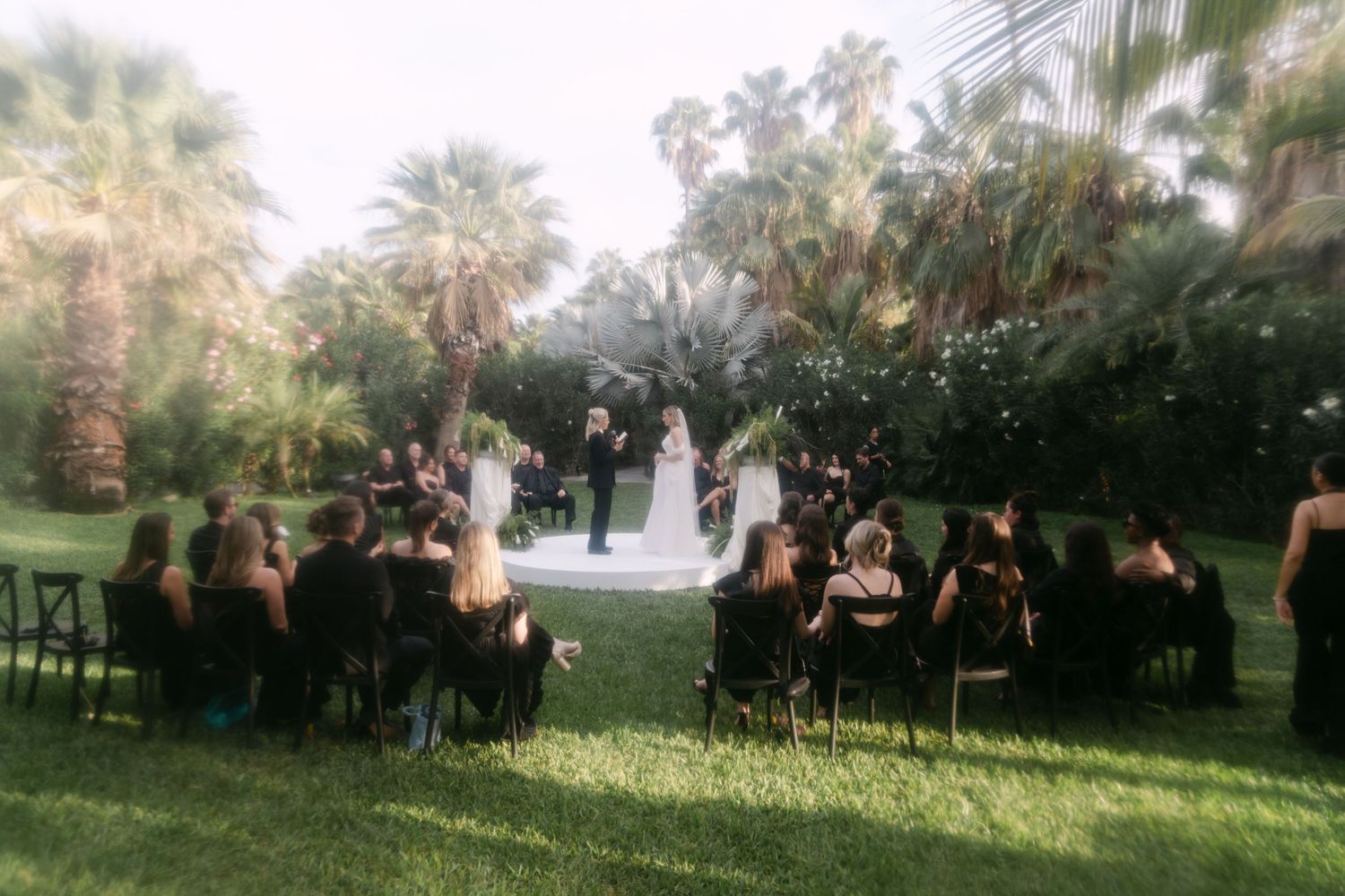Outdoor wedding ceremony on green lawn with guests in black attire surrounded by palm trees.