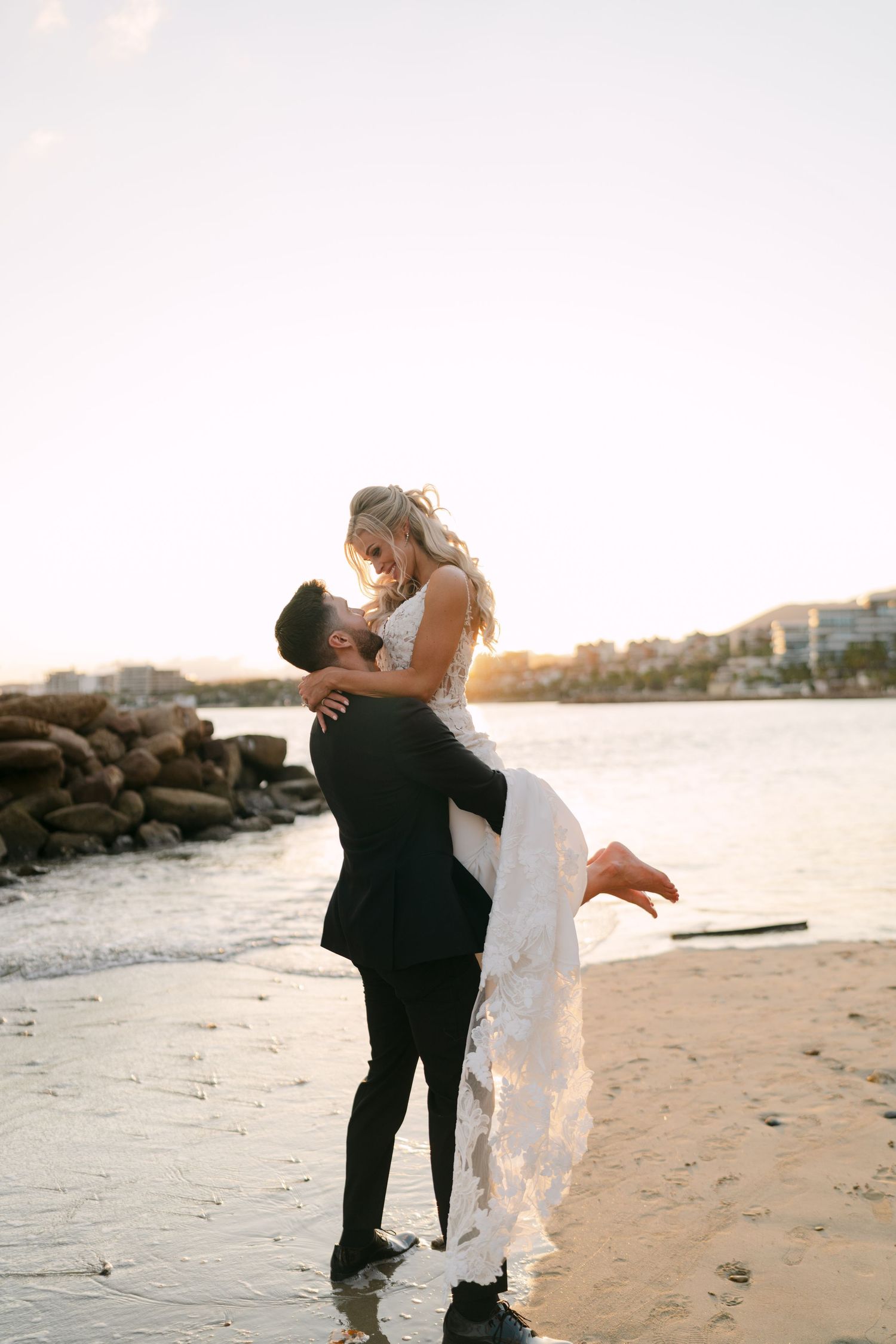 Groom lifts bride in white lace gown on a beach at golden sunset by the water.
