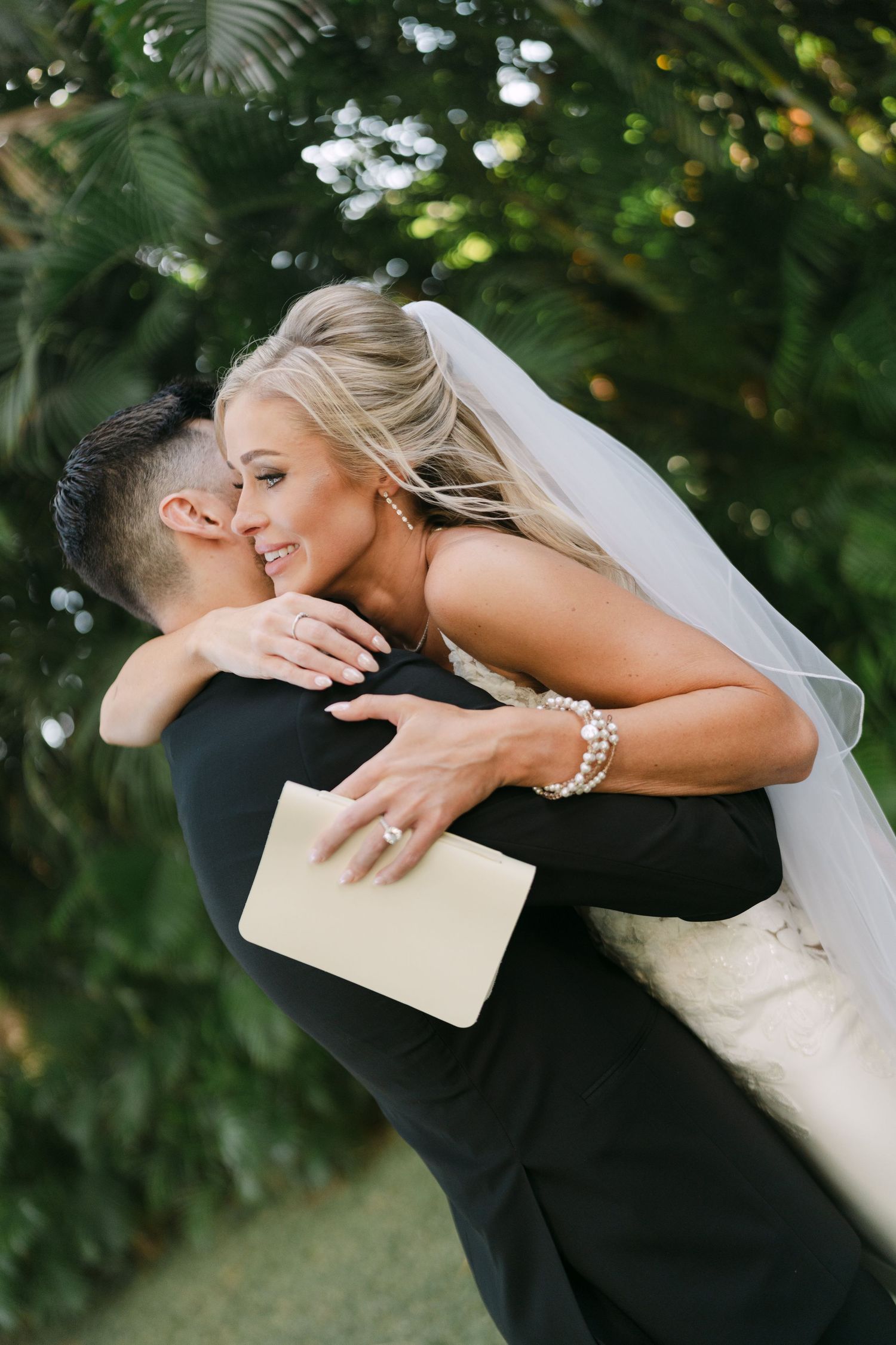 Groom lifting smiling bride in white wedding gown and veil in a romantic embrace surrounded by tropical greenery.