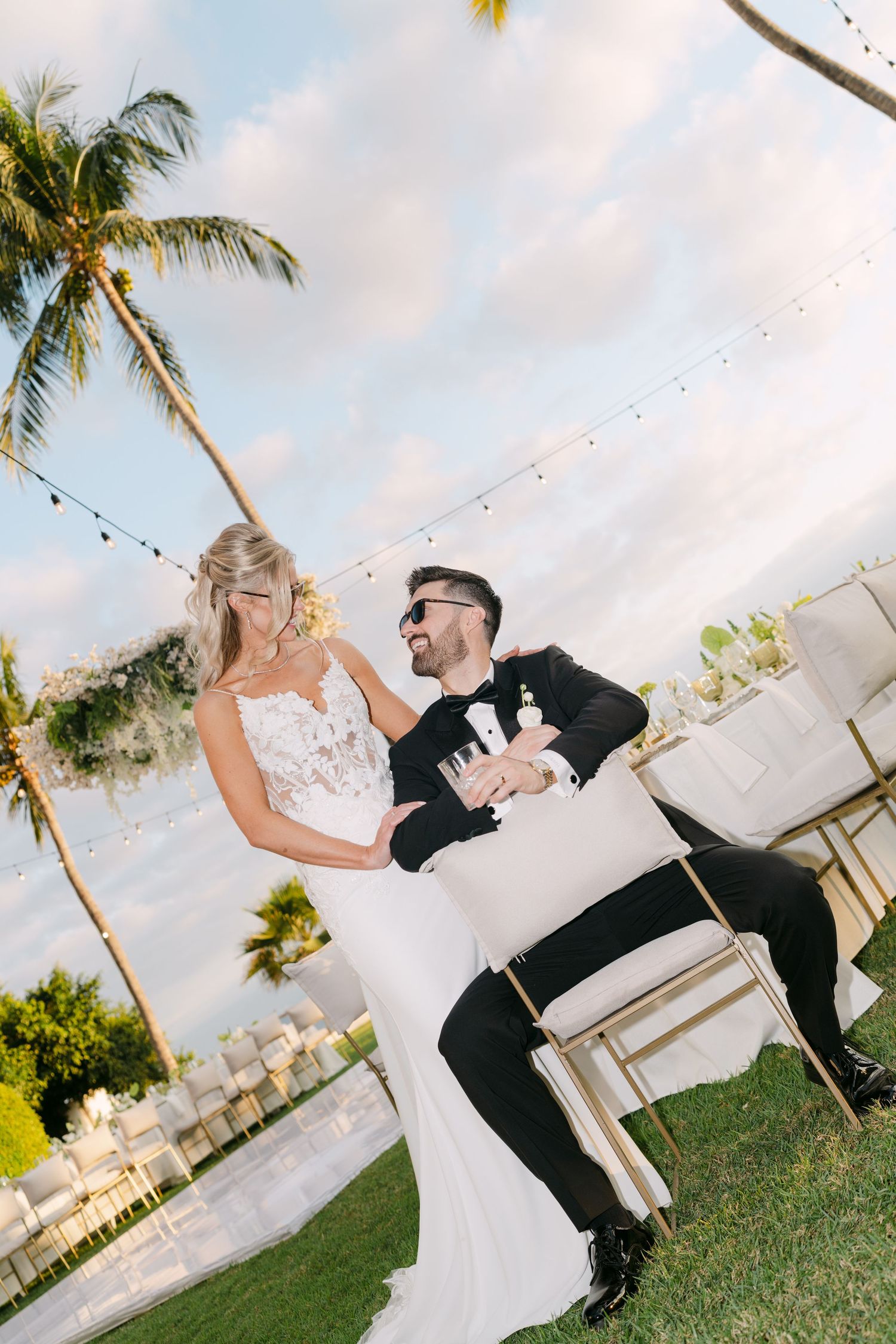 Bride and groom share a joyful moment at an outdoor tropical wedding reception with palm trees and string lights.