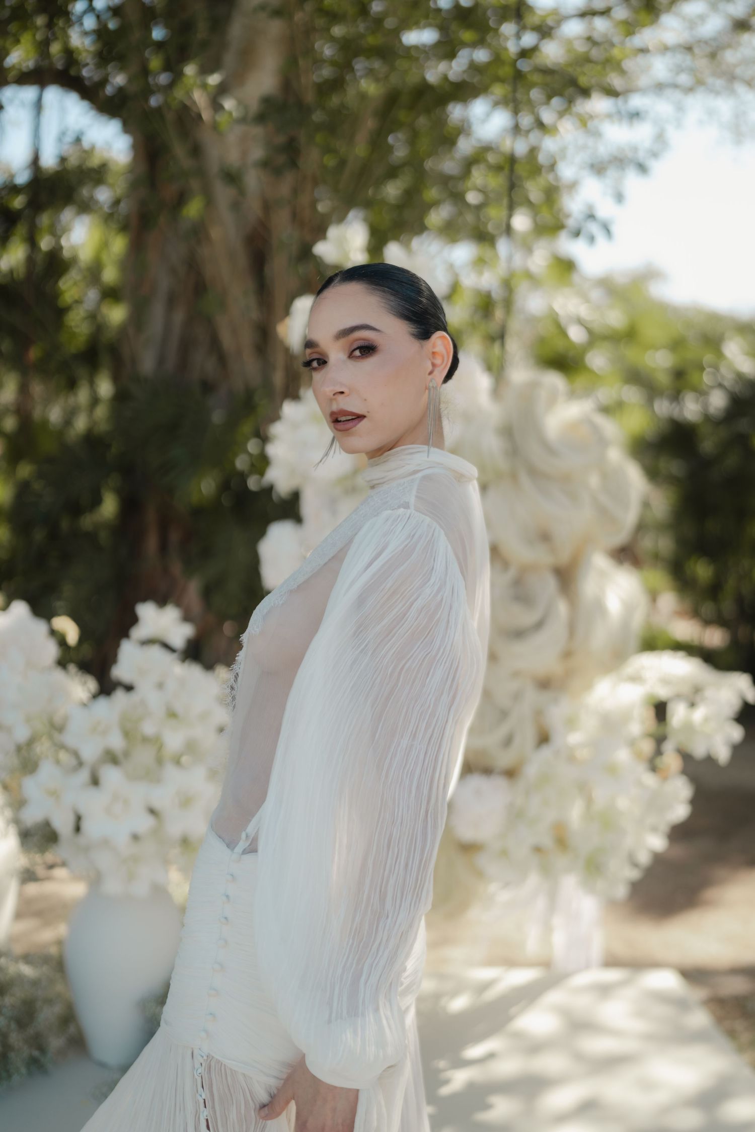 Elegant bride in white sheer long-sleeve gown with fringe detail poses among white floral arrangements outdoors.