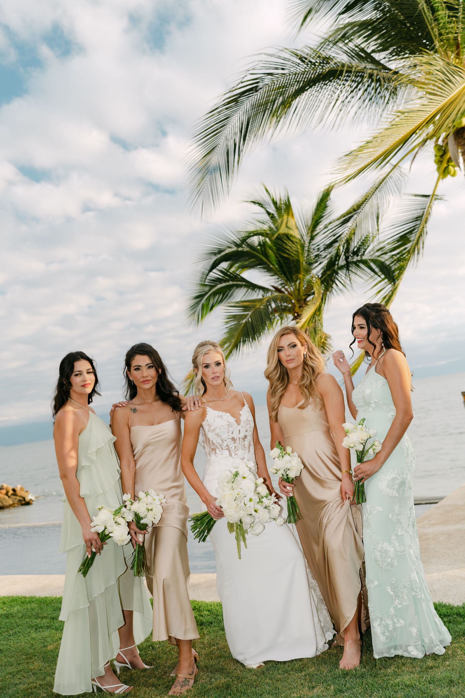 Bride and four bridesmaids in champagne and mint gowns holding white bouquets near a tropical beach palm.