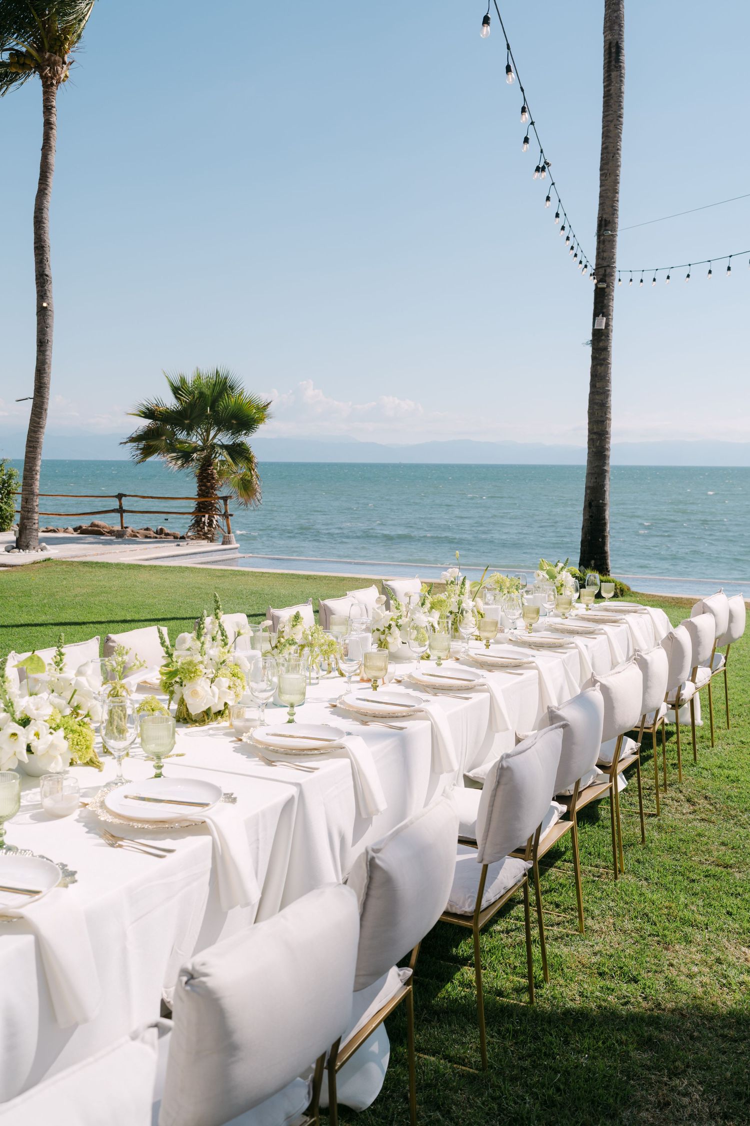 Elegant outdoor wedding reception table set with white linens, gold accents, and white floral centerpieces overlooking the ocean.