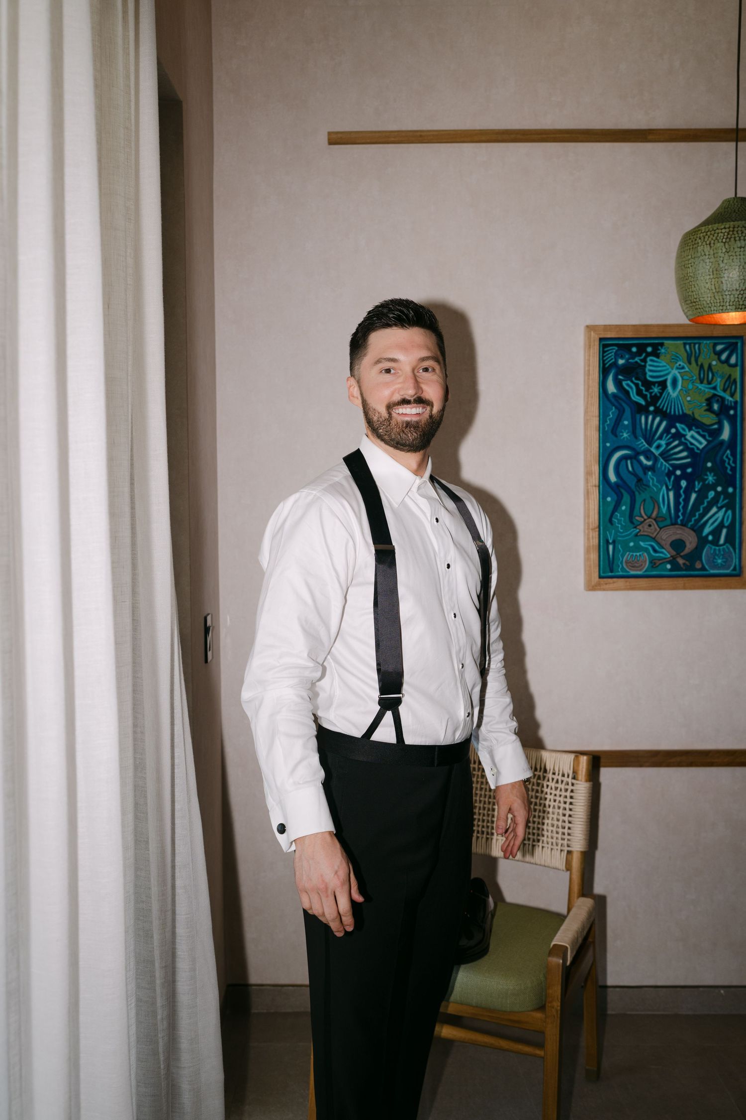 Smiling bearded groom in white dress shirt with black suspenders posing in a stylishly decorated hotel room.