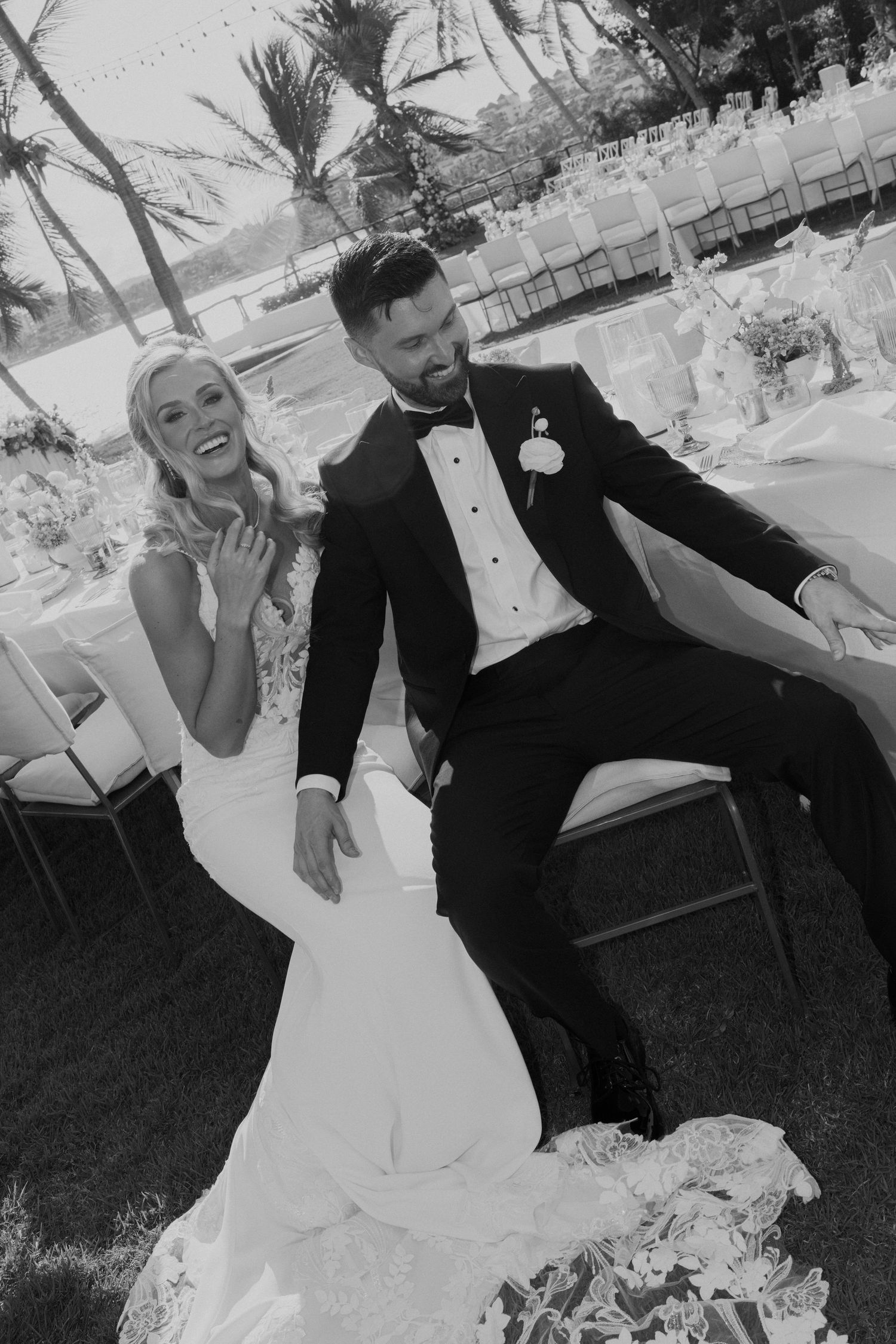 Black and white photo of laughing bride in fitted gown and bearded groom in tuxedo seated at outdoor reception.