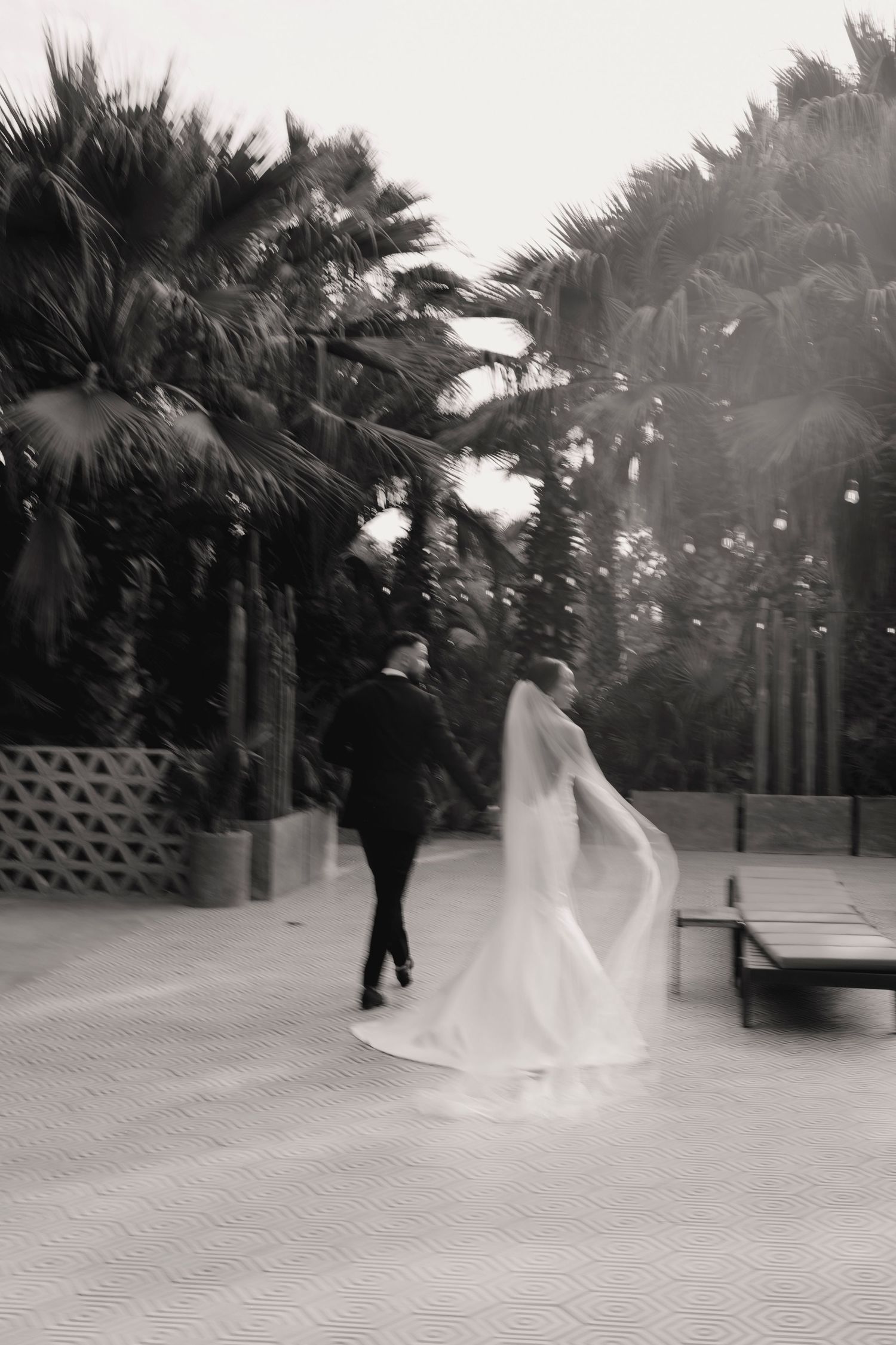 Black and white motion blur photo of bride and groom walking among tropical palm trees together.