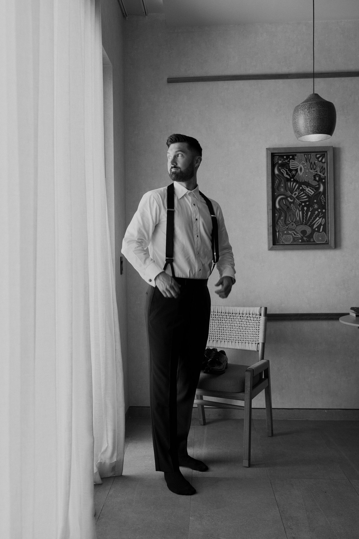 Black and white portrait of groom in white shirt and suspenders gazing out a window in a modern hotel room.