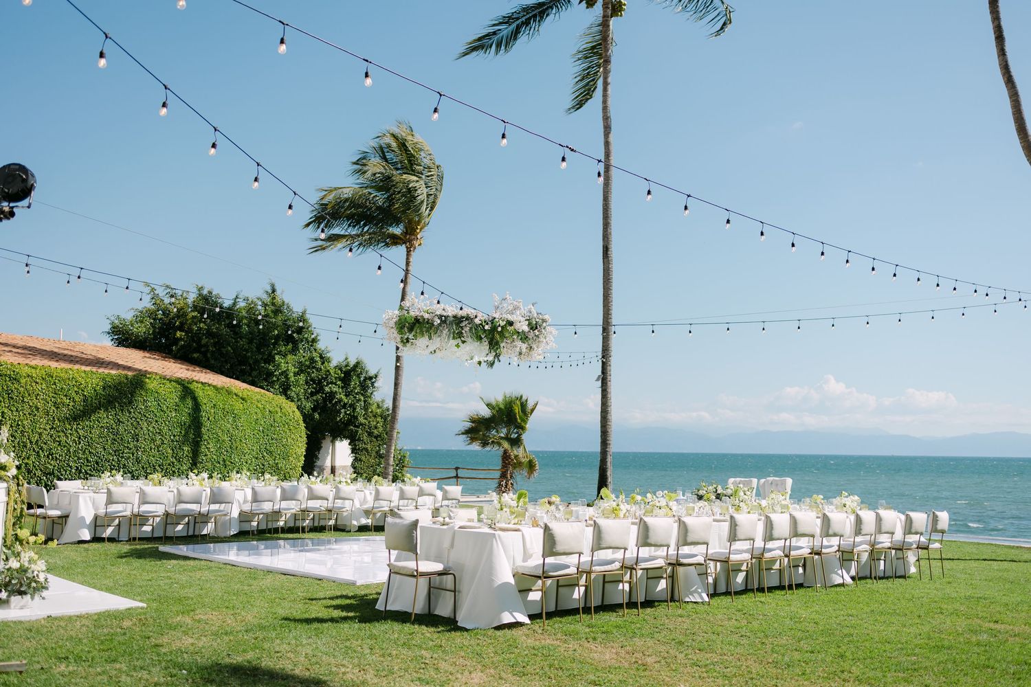 Outdoor beachside wedding ceremony setup with white chairs, string lights, and palm trees under a clear blue sky.