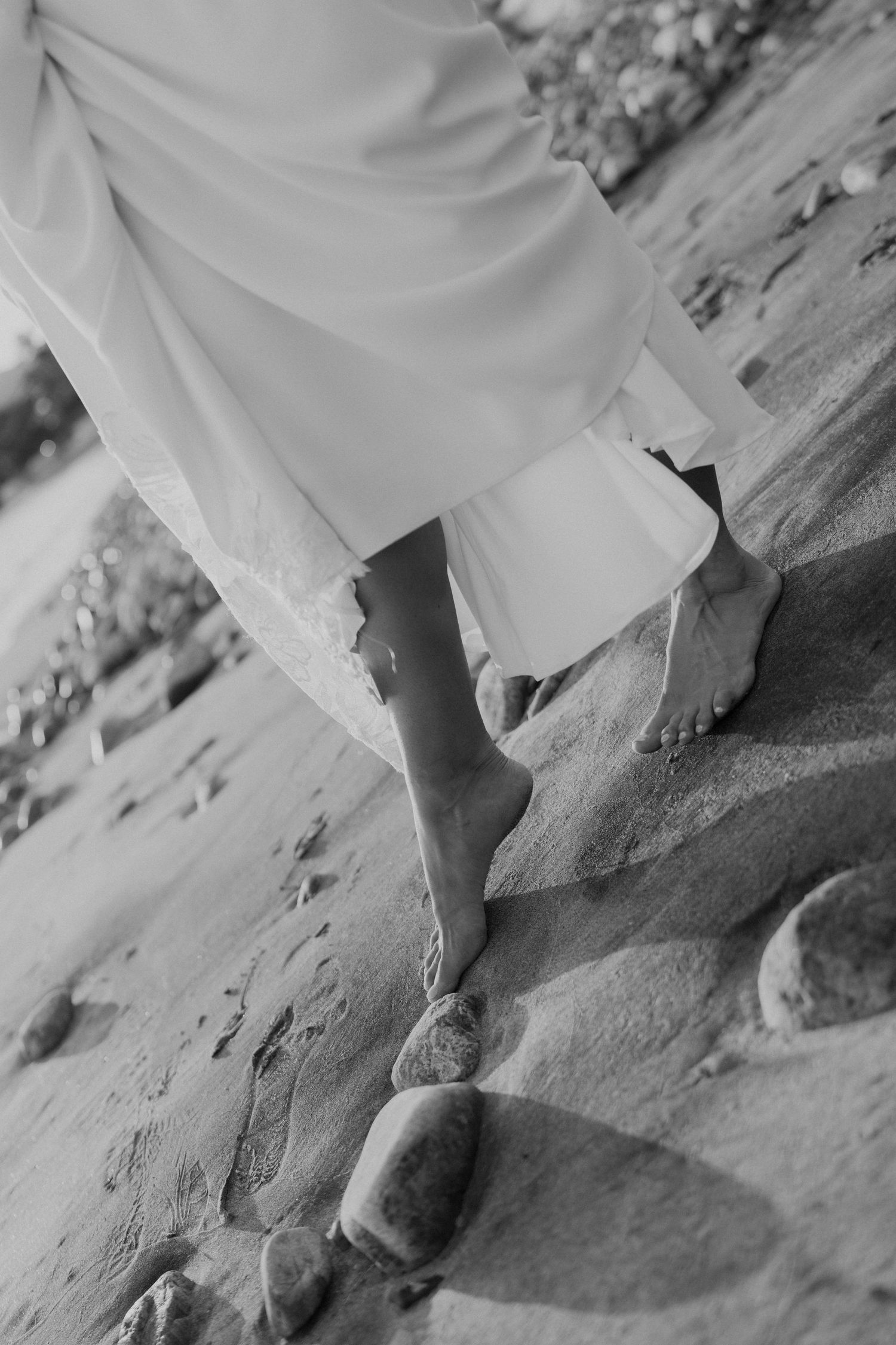 Barefoot bride walking on sandy beach rocks in flowing white wedding dress, black and white photography.