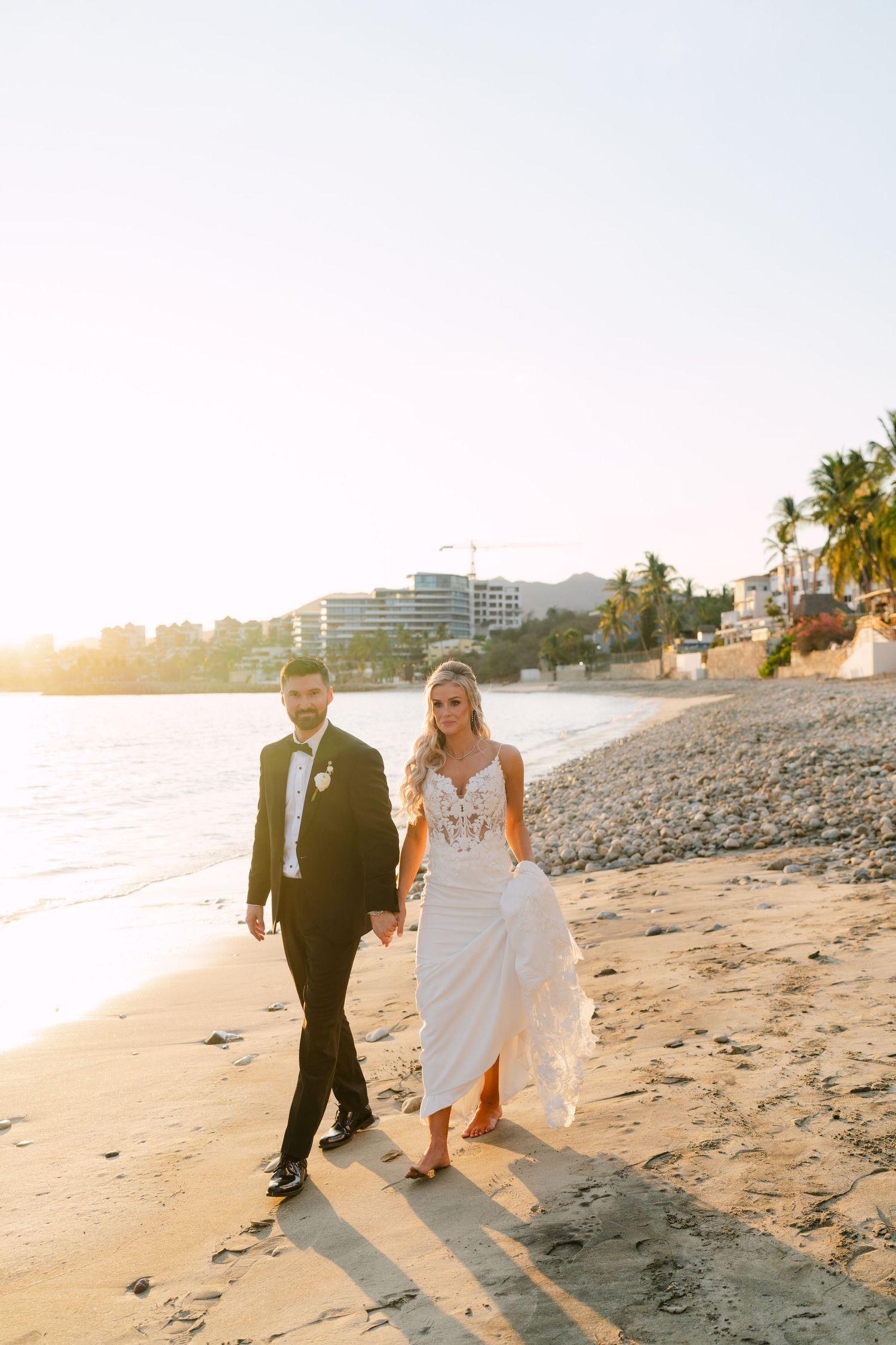 Bride and groom walk hand in hand along a sandy beach at golden hour during their wedding day.
