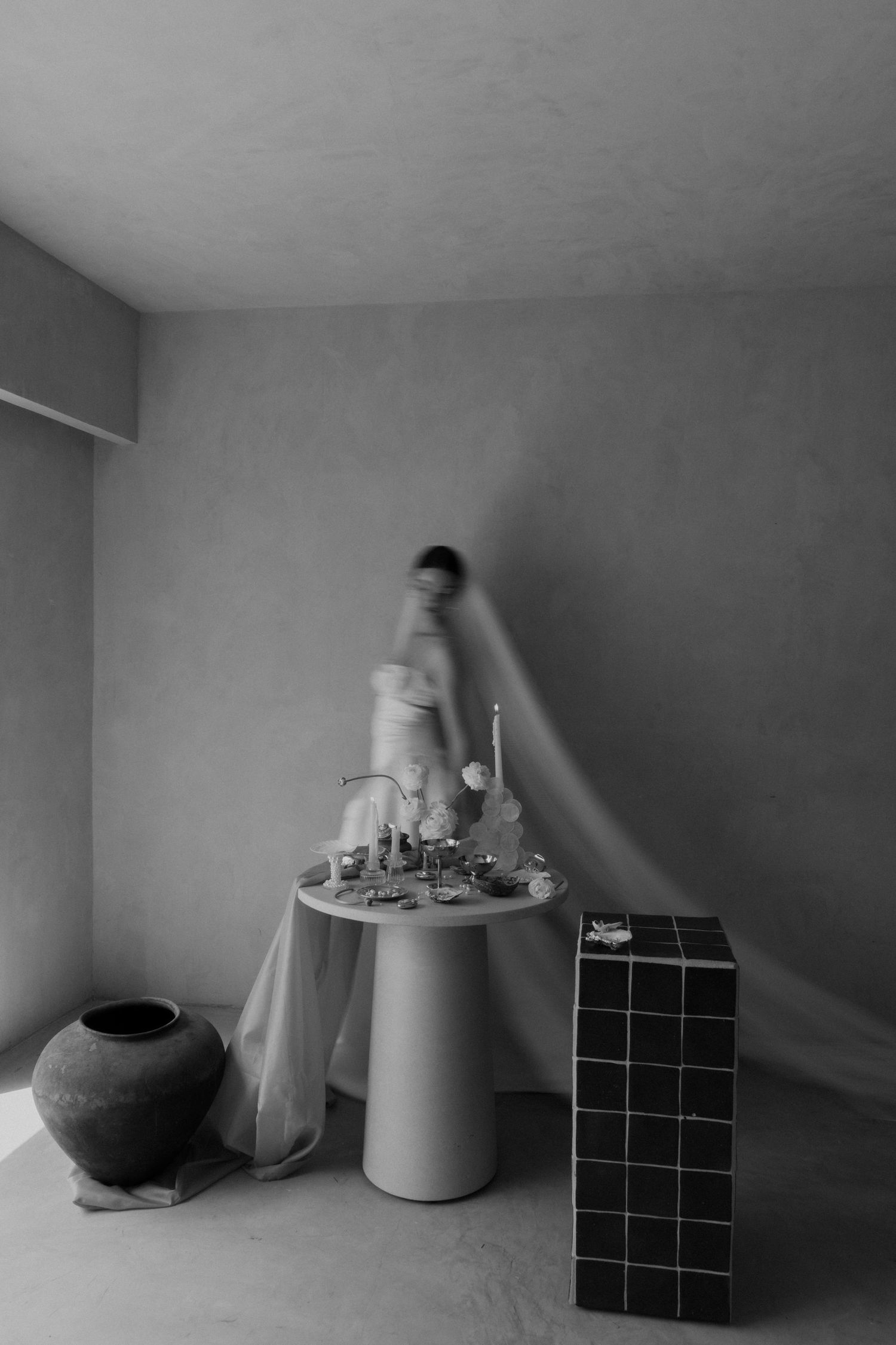 Blurred bride in white gown moving past a styled table with candles and florals in a minimalist gray room.