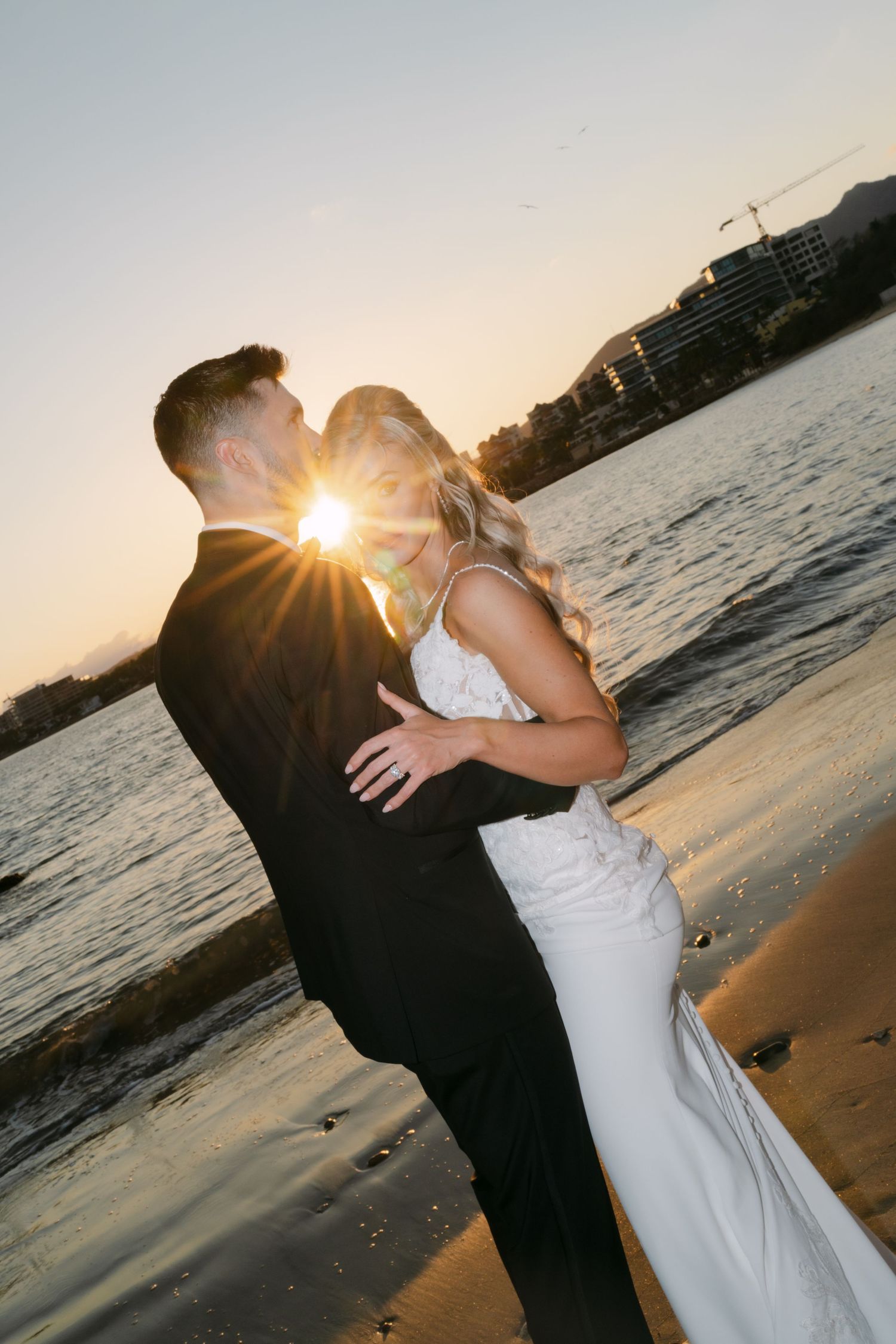Bride and groom share a romantic sunset kiss on the beach with golden light glowing between them.