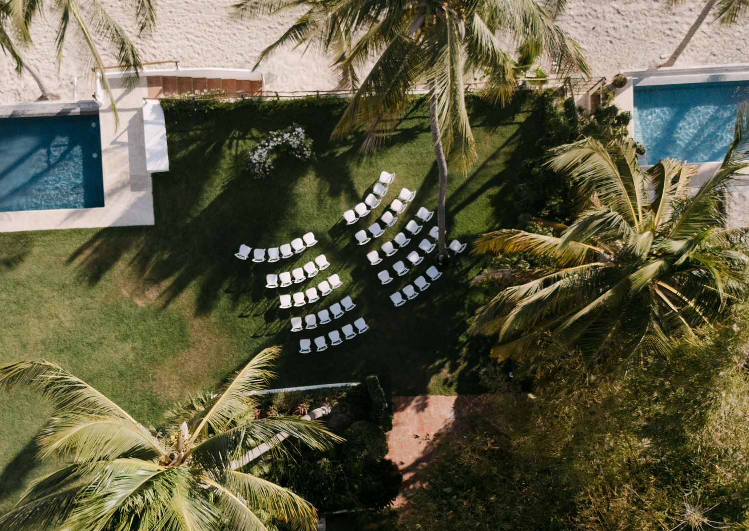 Aerial view of white wedding ceremony chairs arranged in curved rows on green lawn surrounded by palm trees.