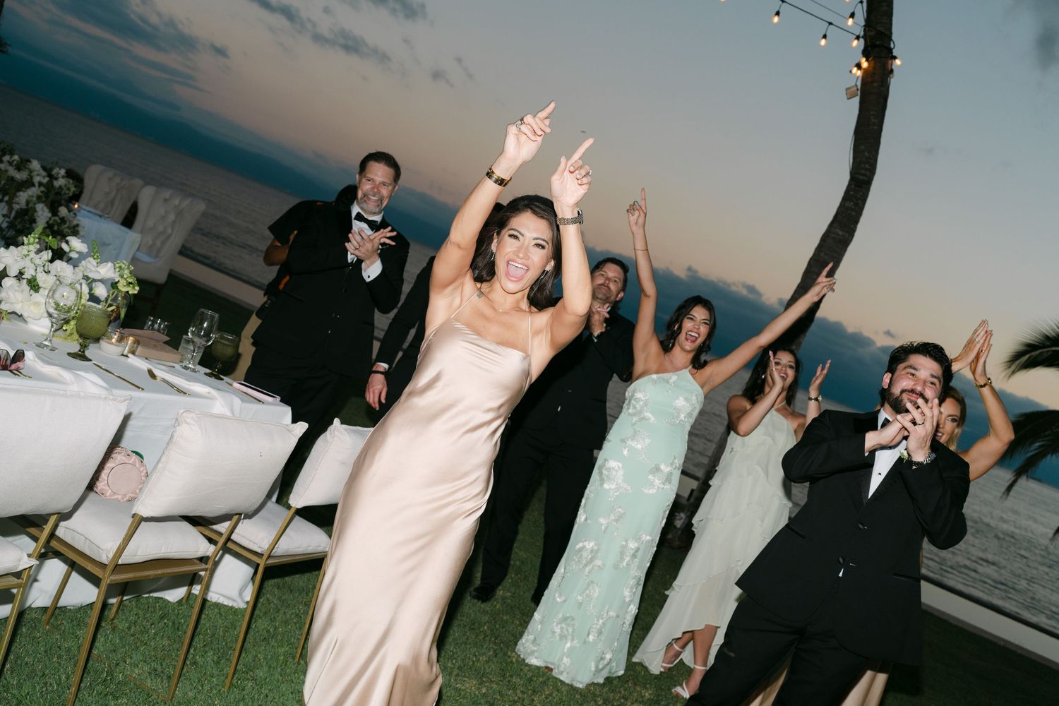 Joyful wedding reception celebration with bride in champagne gown dancing outdoors by the ocean at dusk.
