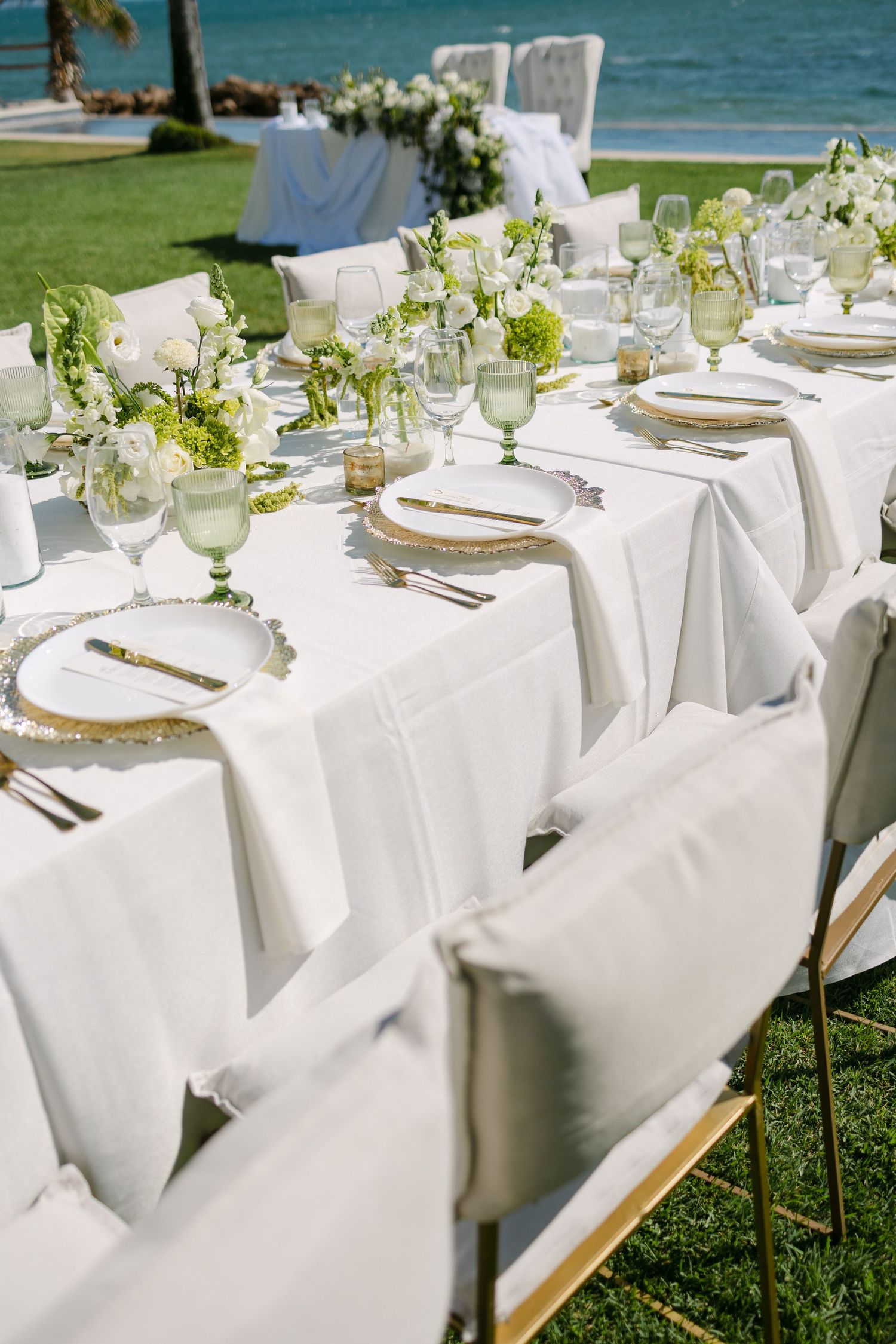 Elegant outdoor wedding reception table set with white linens, green florals, gold chargers, and glassware.