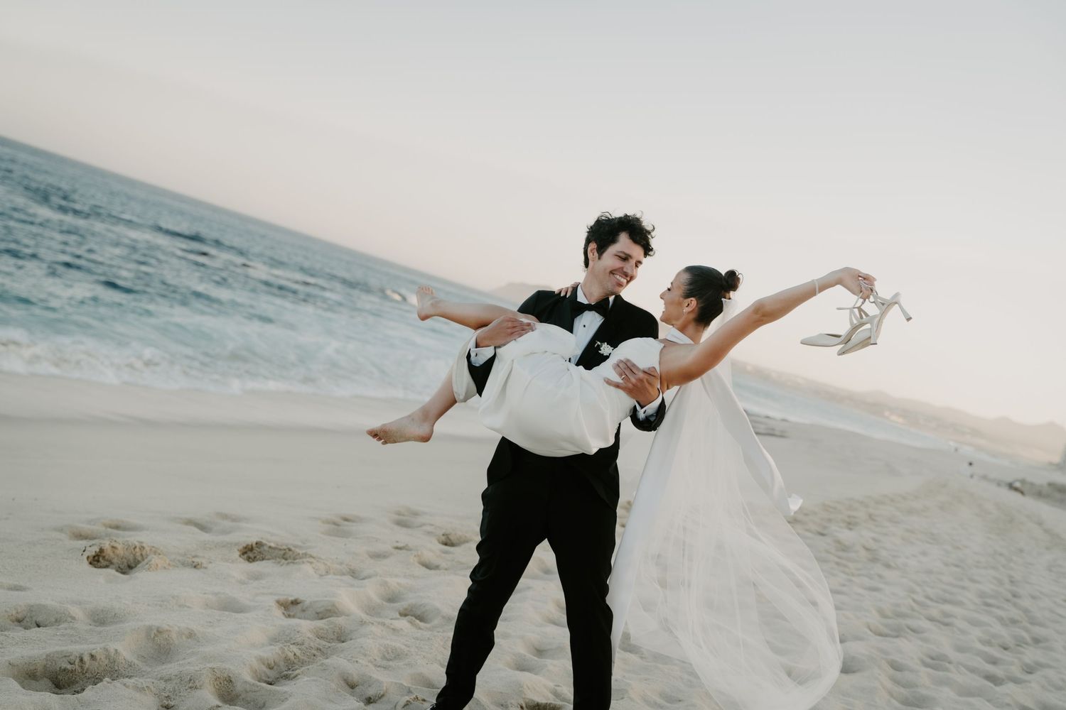 Groom in black tuxedo carries laughing bride in white gown on sandy beach at sunset.