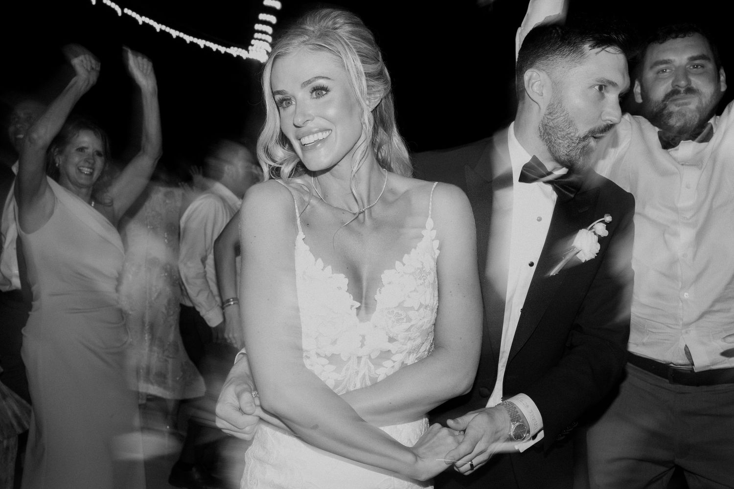 Black and white photo of bride and groom dancing joyfully with guests at their wedding reception.