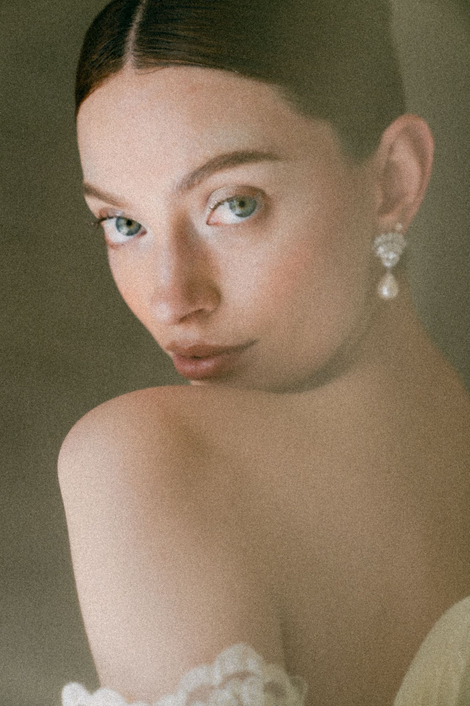 Close-up portrait of a young woman with blue eyes wearing elegant pearl drop earrings and an off-shoulder white lace dress.