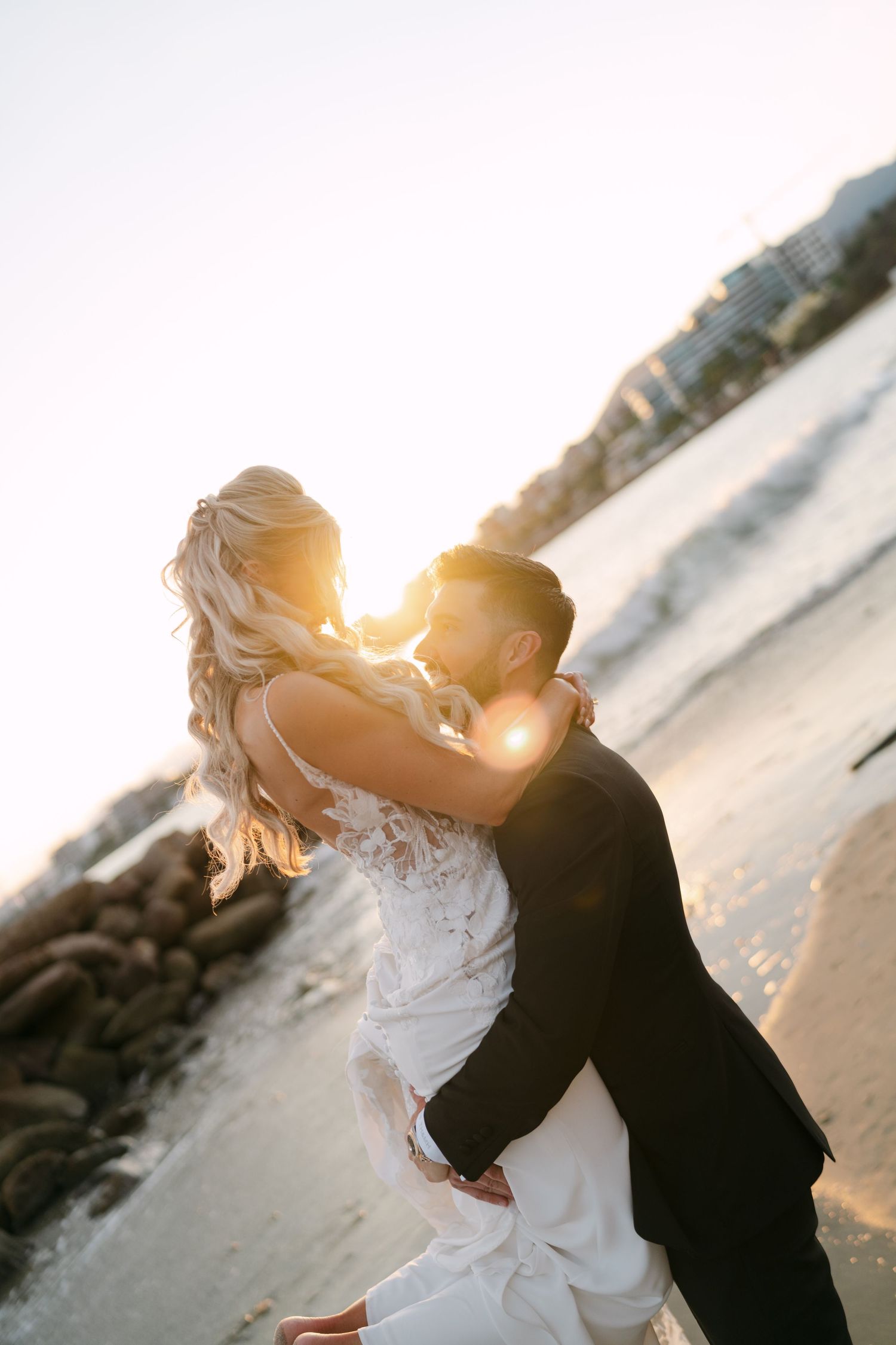 Groom lifts bride as they share a kiss on the beach with stunning golden sunset light behind them.