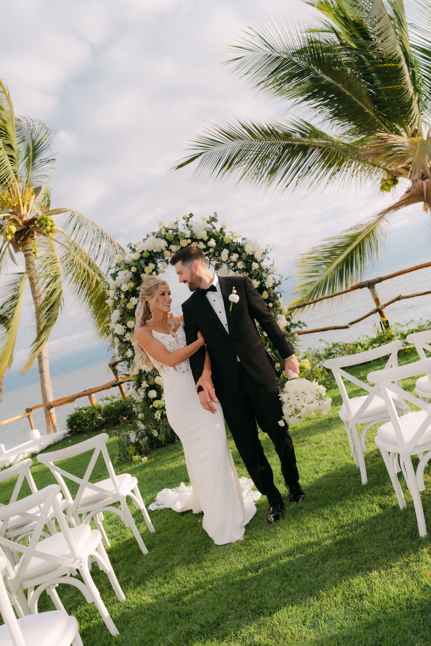 Bride and groom standing at floral arch ceremony altar surrounded by palm trees and white chairs overlooking the ocean.