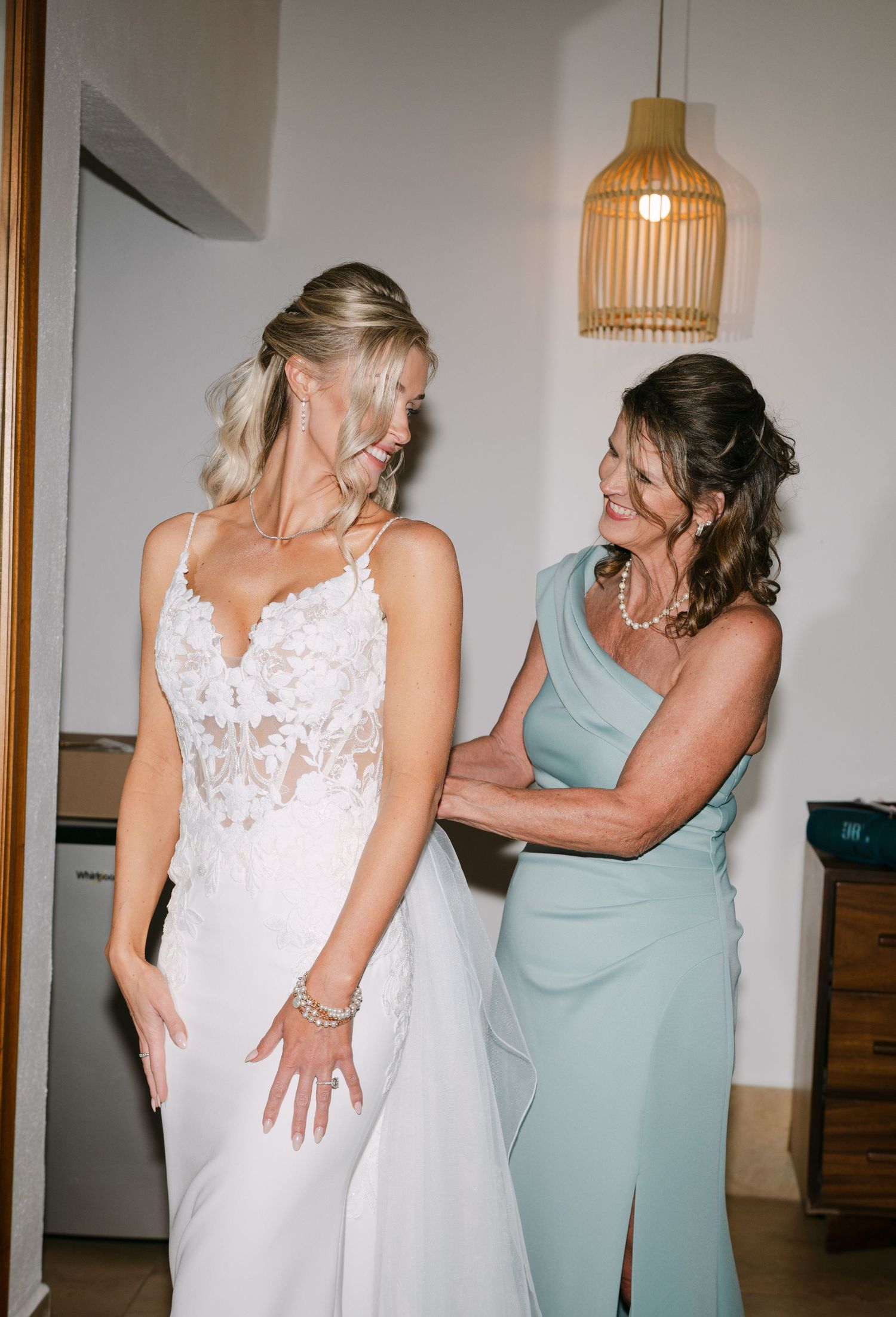 Mother helping bride in lace wedding gown get dressed under warm pendant light before the ceremony.