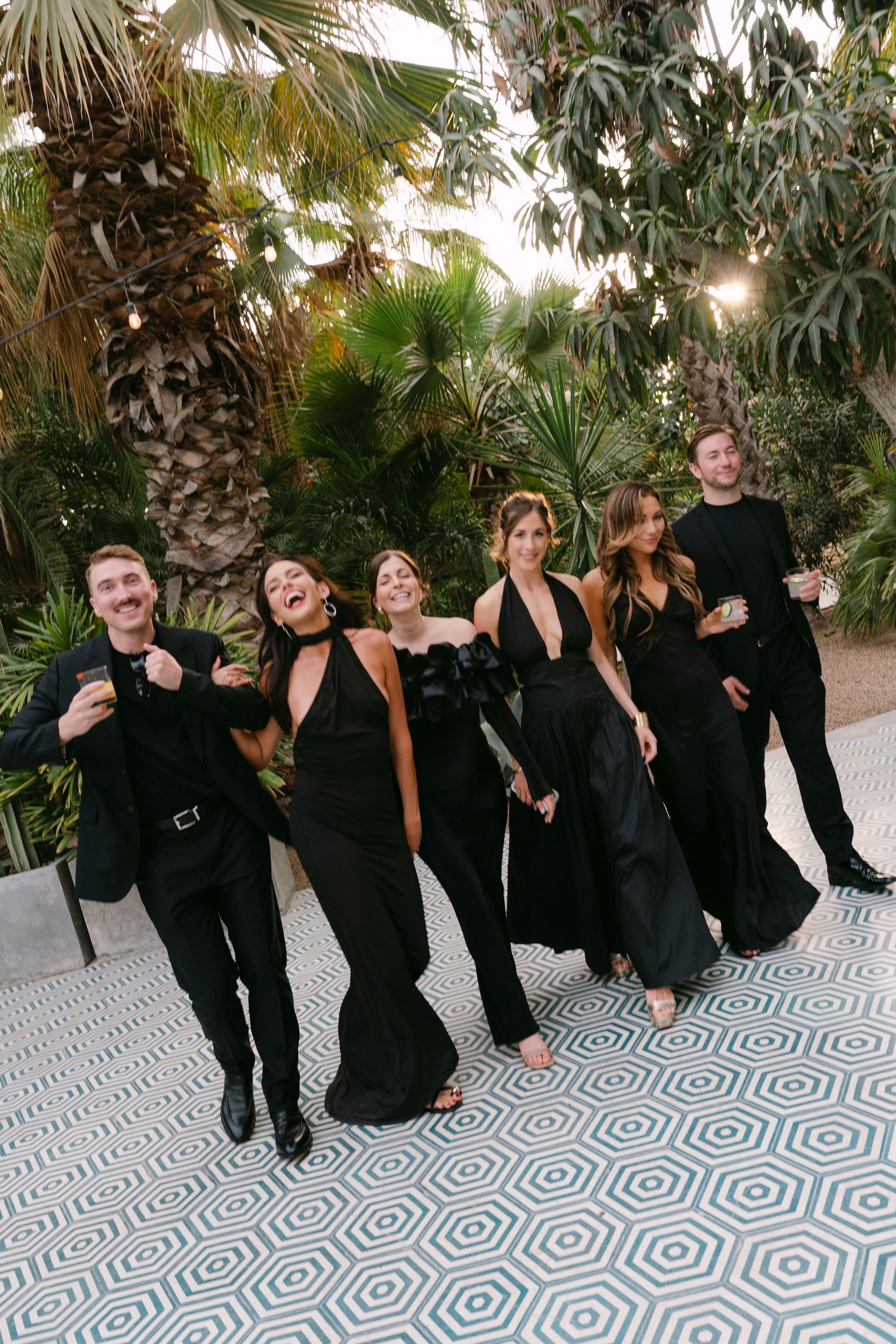 Wedding party in matching black formal attire poses joyfully on geometric patterned floor outdoors.