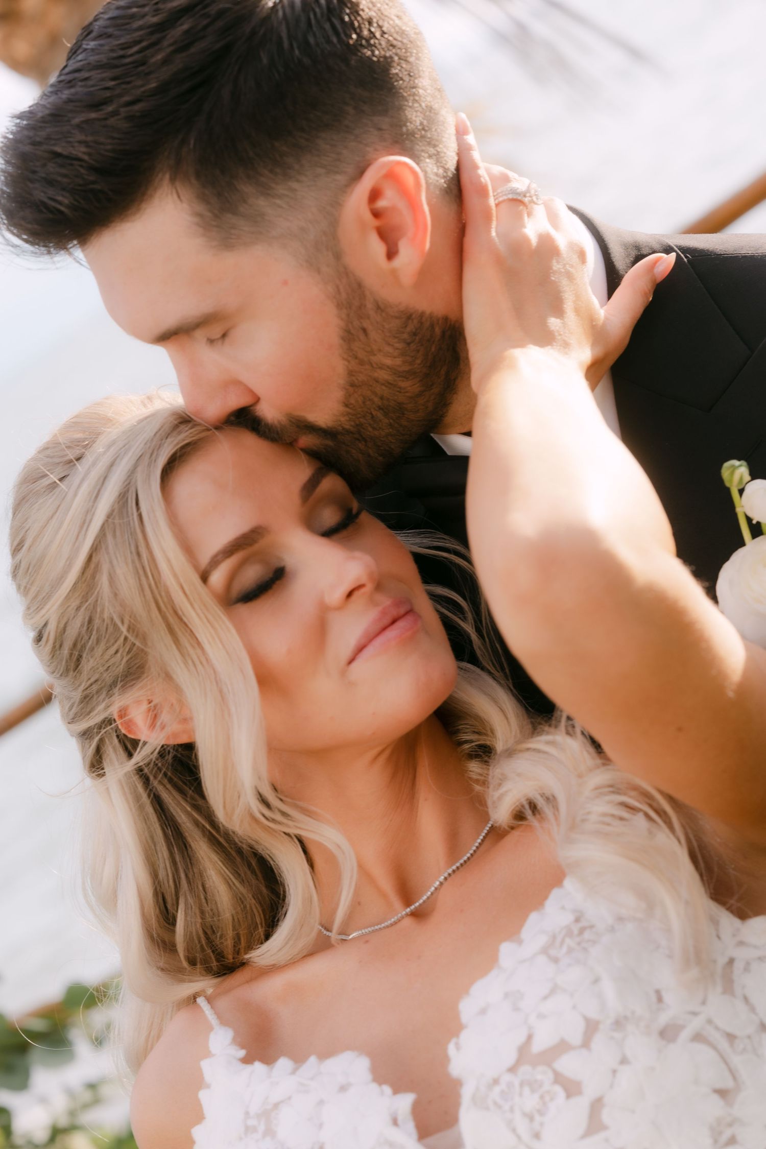 Groom tenderly kissing bride on the temple as she closes her eyes in a romantic outdoor wedding portrait.